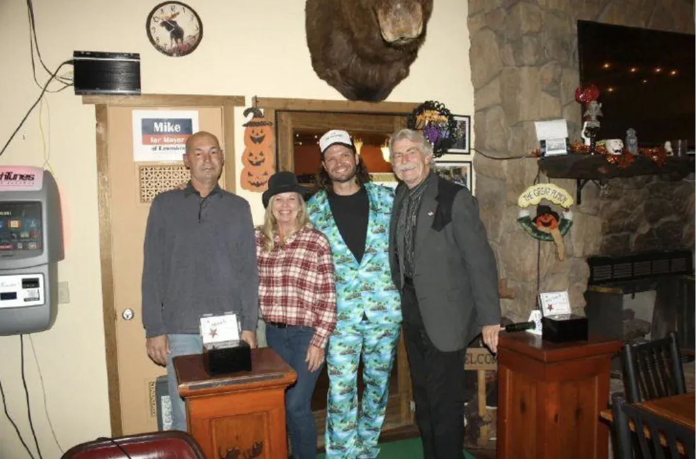 Four people pose indoors, in front of a doorway and stone fireplace; one wears a blue suit.