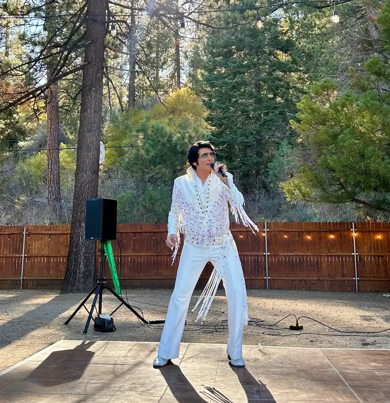 Man dressed as Elvis singing outdoors, white sequin suit, microphone, stage, trees, wooden fence.
