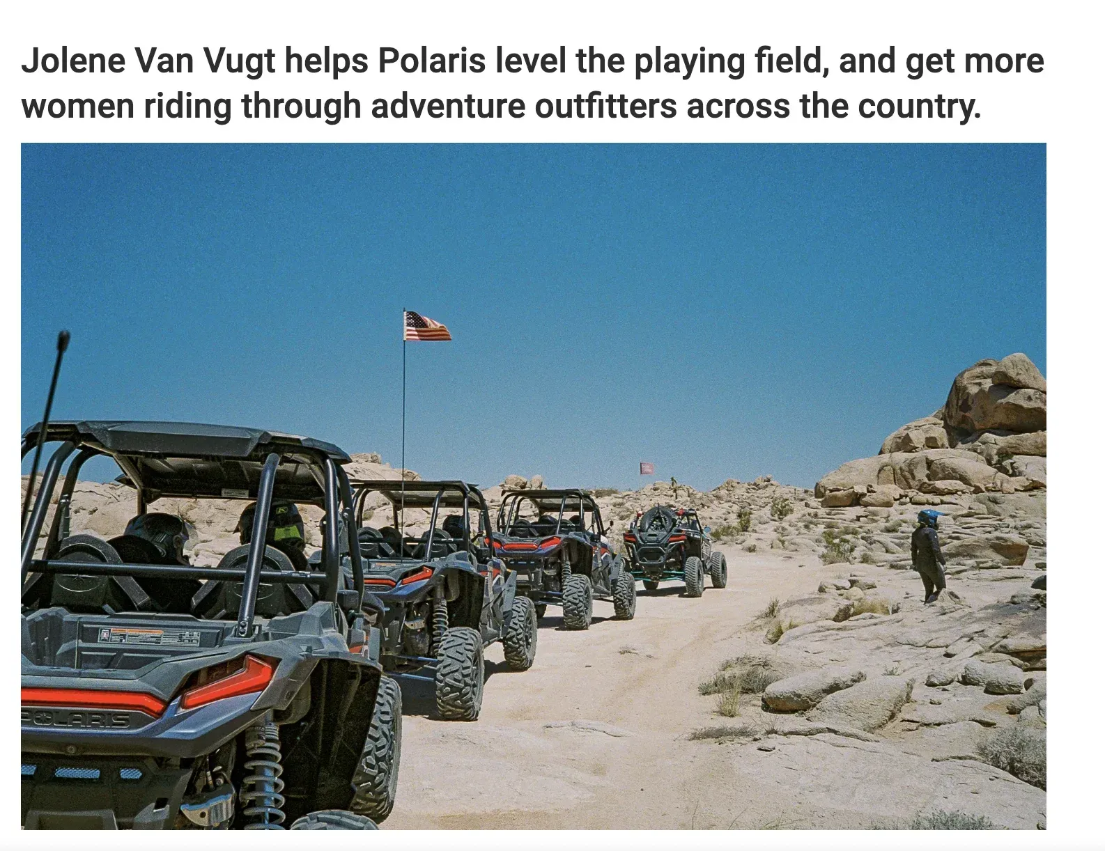 A line of off-road vehicles drives on a dirt path in the desert. A woman stands to the side.