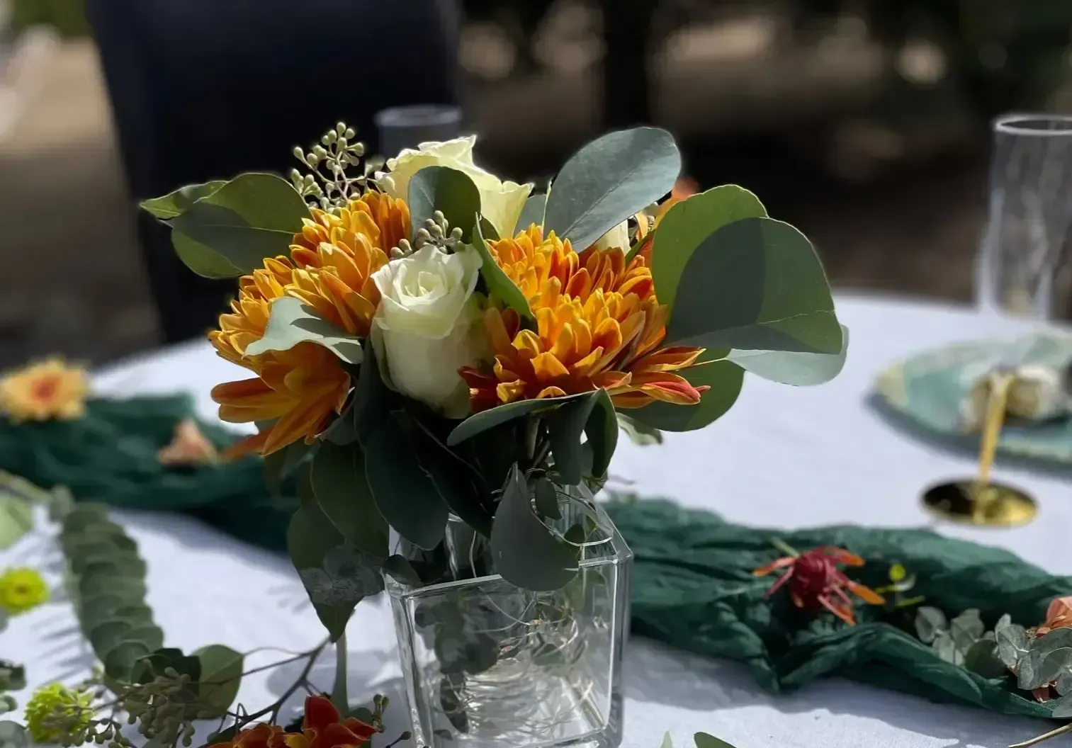 Close-up of a flower arrangement in a clear glass vase on a table. Yellow and orange blooms with green foliage.