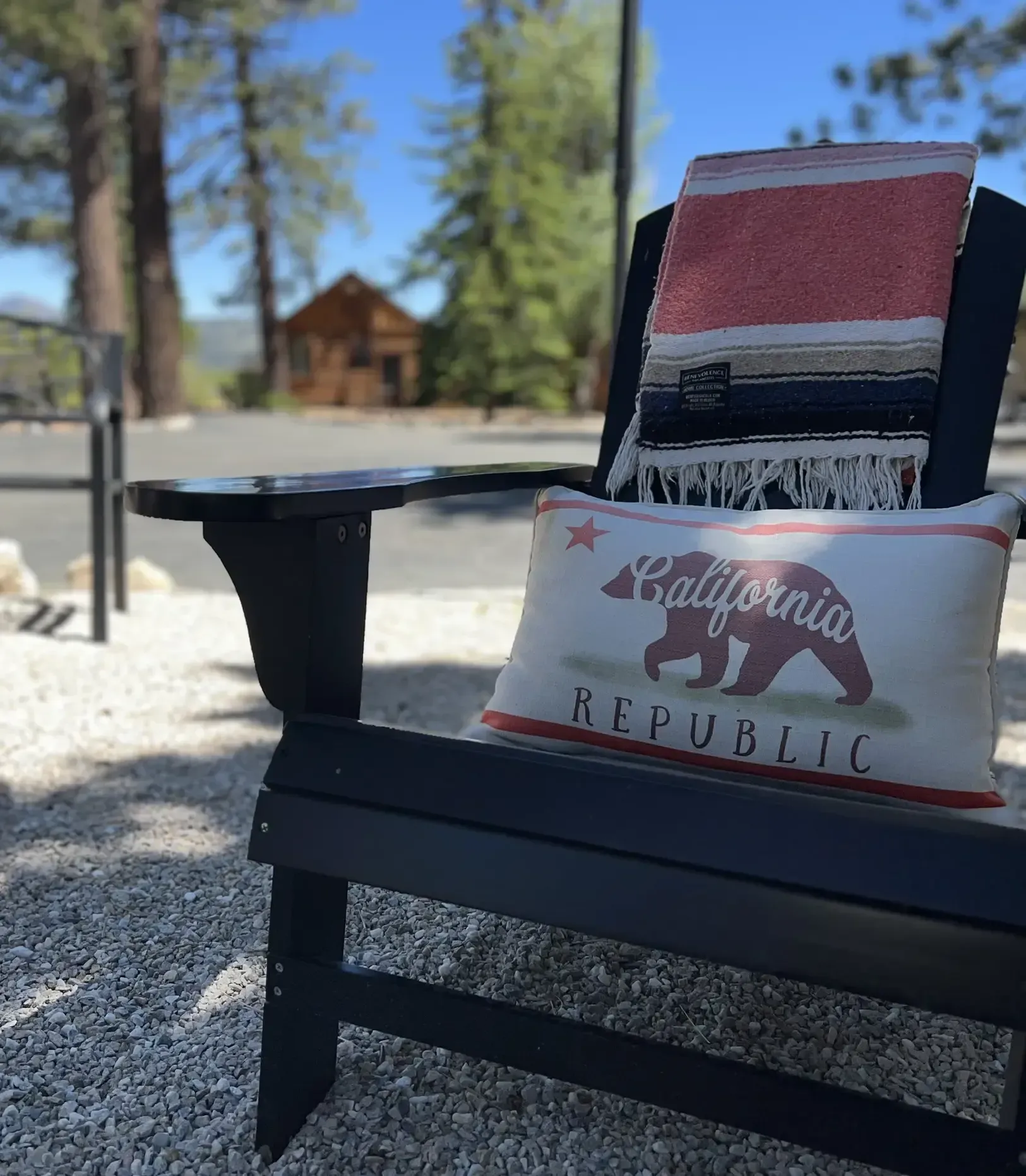 Black Adirondack chair with pillow and blanket in front of a cabin, sunny outdoor setting.