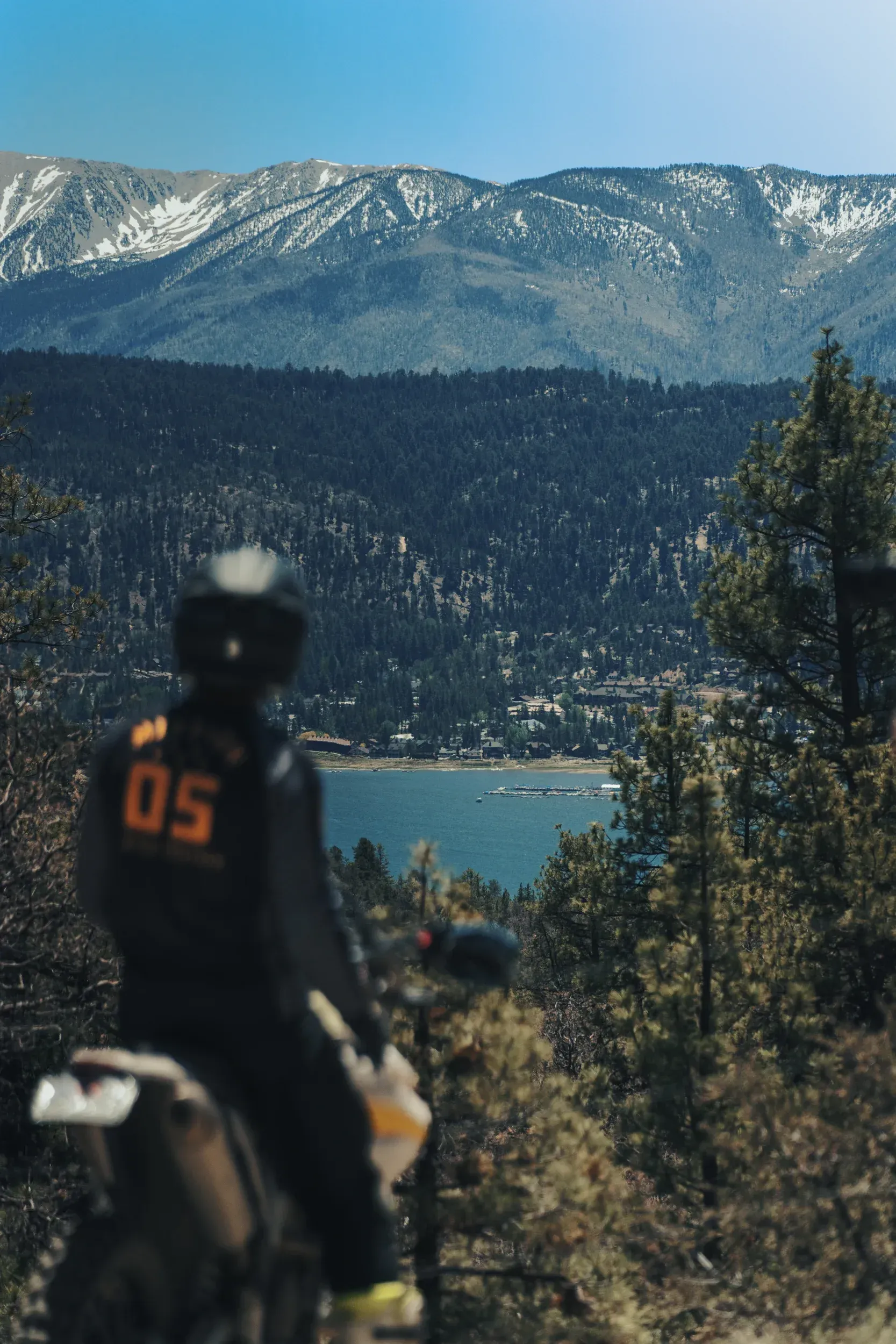 Motorcyclist overlooking a lake and mountain range. Snow-capped peaks, evergreen forest, blue water.