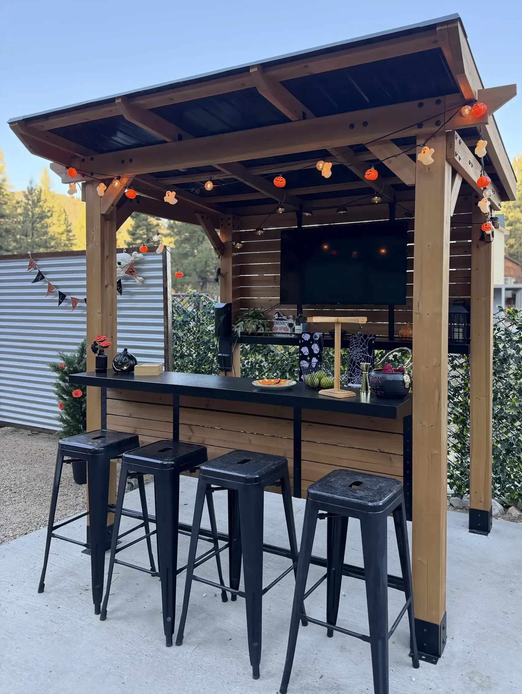 Outdoor bar with black stools and a TV, beneath a wooden pergola decorated with string lights.
