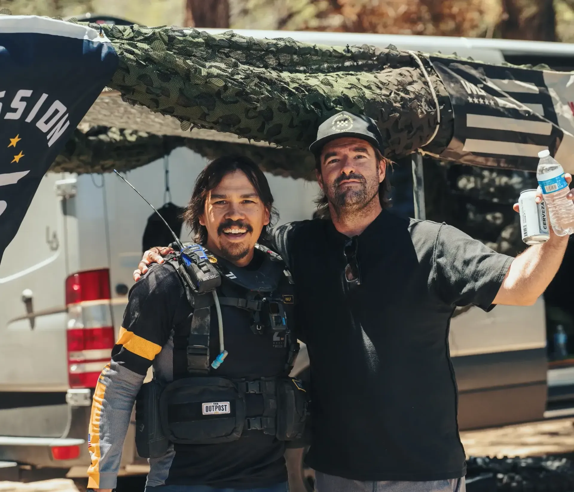 Two men pose in front of a white van with camouflage netting. One holds drinks, the other wears a tactical vest and smiles.