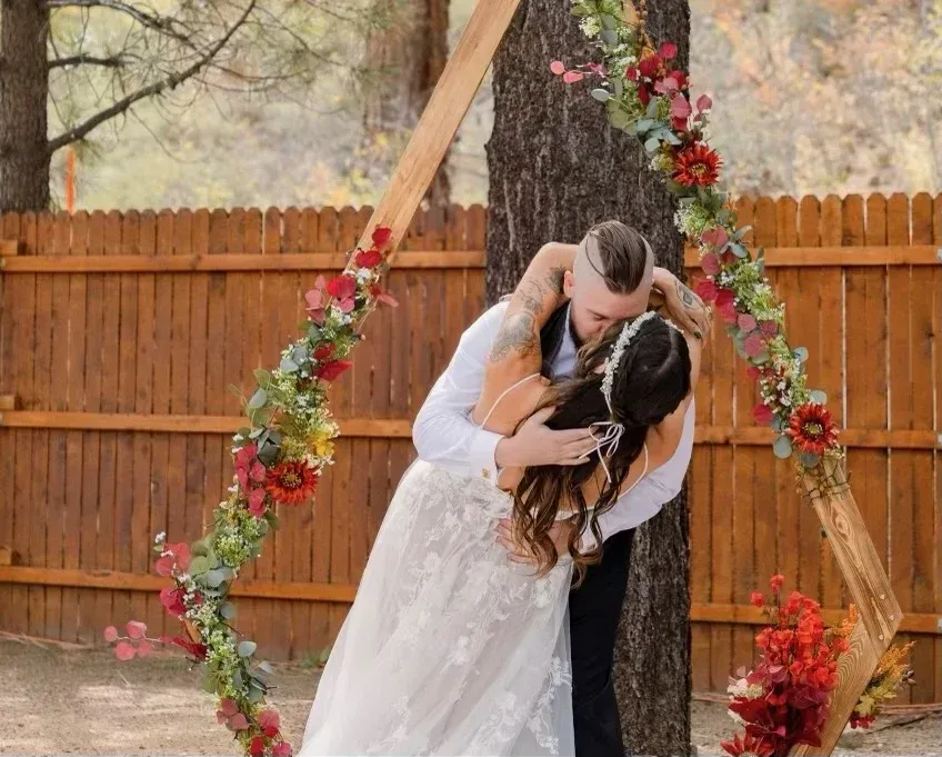 Couple embraces, kissing beneath a wooden wedding arch decorated with red flowers, near a fence.