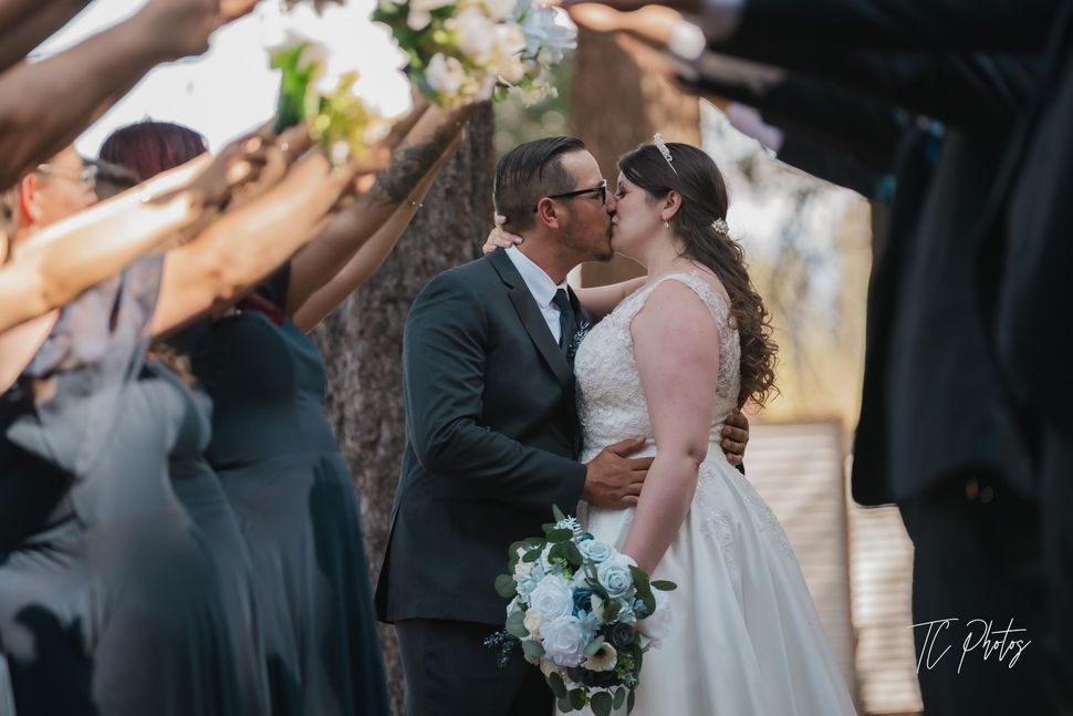 Man in green coat lifts bride in white dress, both smiling, outdoors.