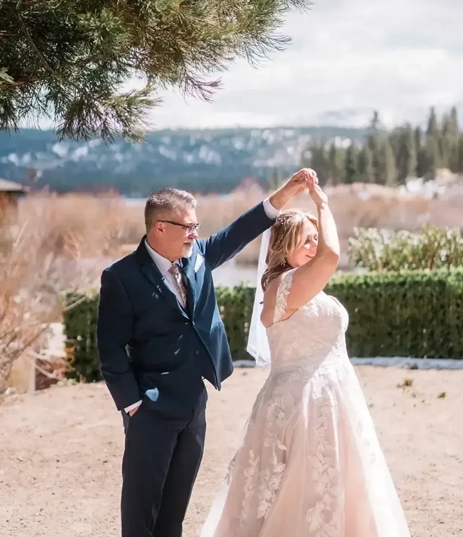 Bride and groom dancing outdoors; man in navy suit, woman in white gown, sunny day, mountains in the background.