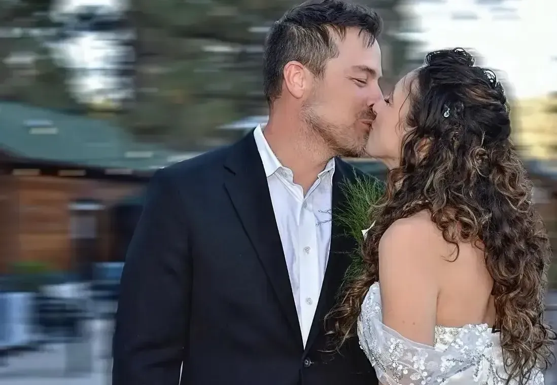 Couple kissing, man in suit, woman in white wedding dress, outdoors by building.