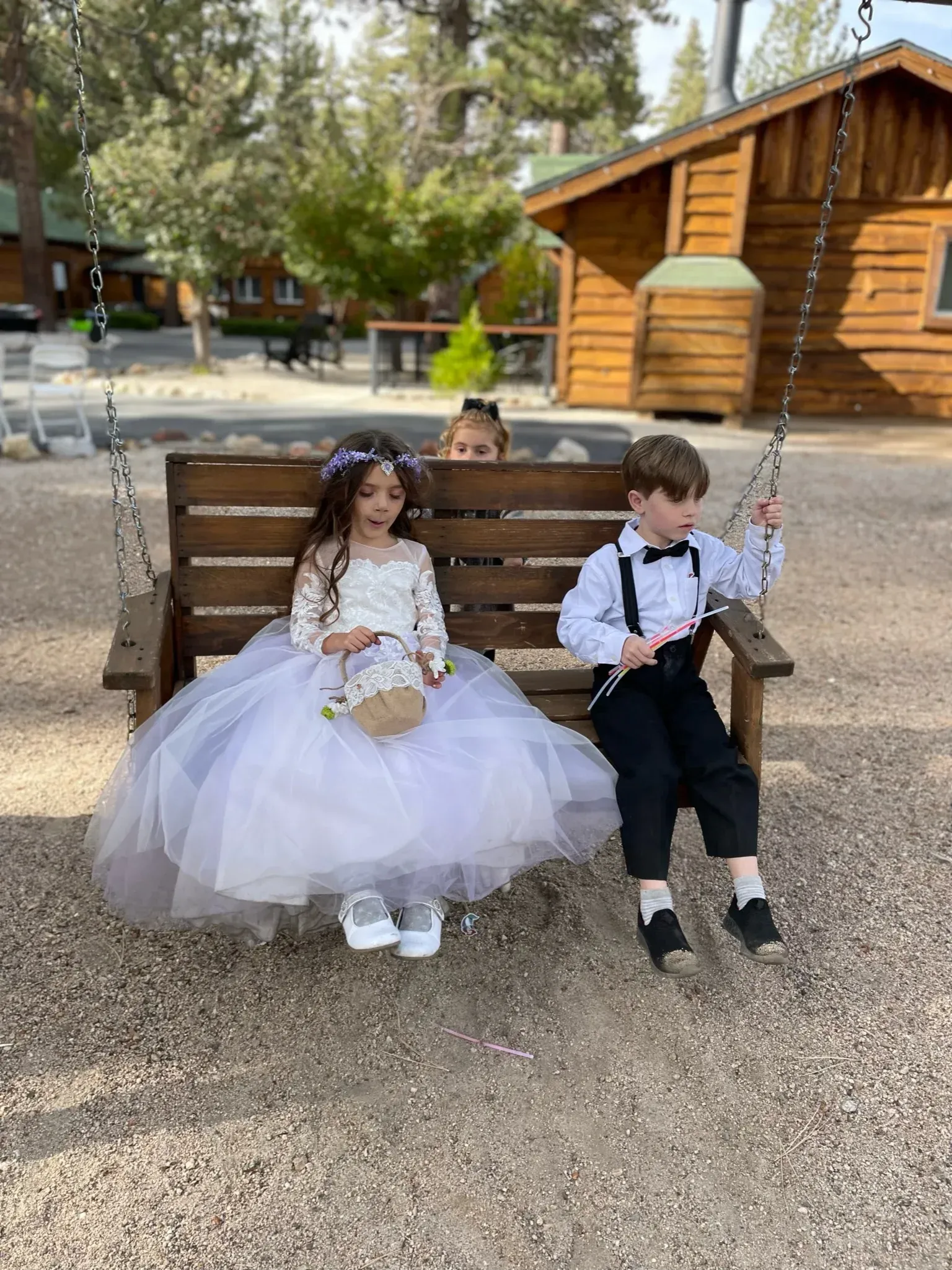 Children on a swing: girl in white dress, boy in bow tie, another child peeking behind. Wooden cabin in background.