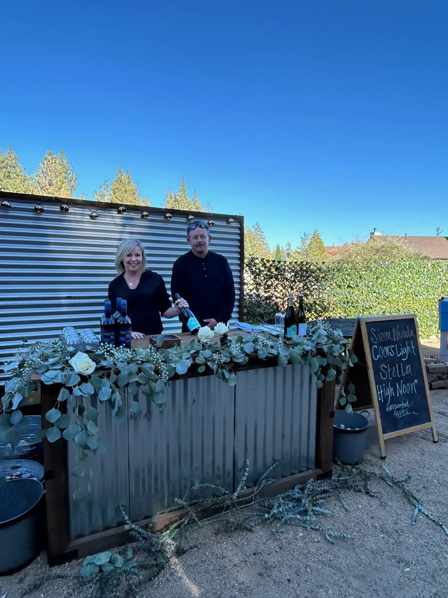 Two people behind a decorated outdoor bar. Blue sky, greenery, and galvanized metal.