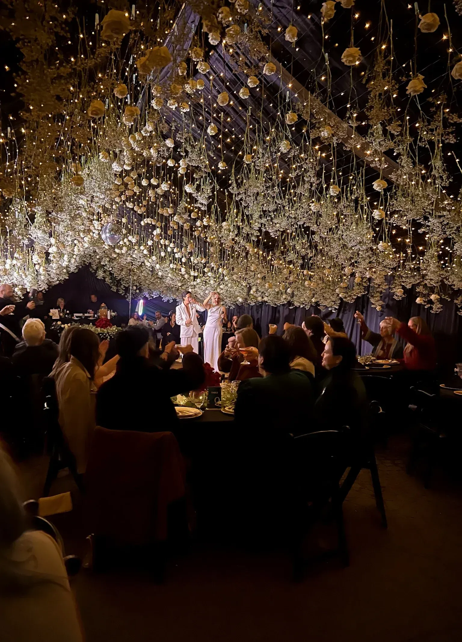 Wedding reception with overhead floral decorations, guests seated at tables, and figures in the background.
