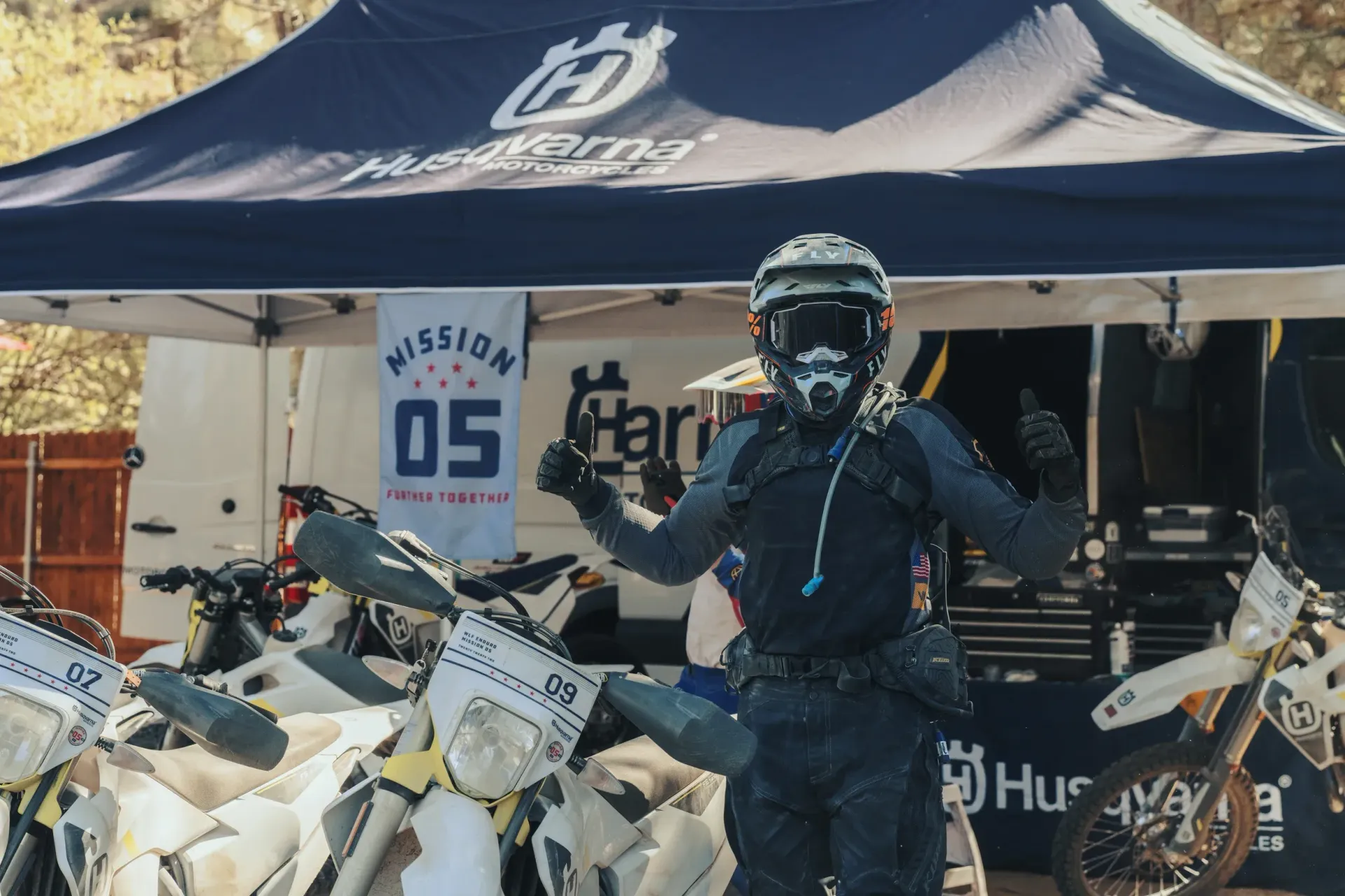 Motorcyclist in helmet and gear, giving thumbs-up, in front of Husqvarna tent and bikes.
