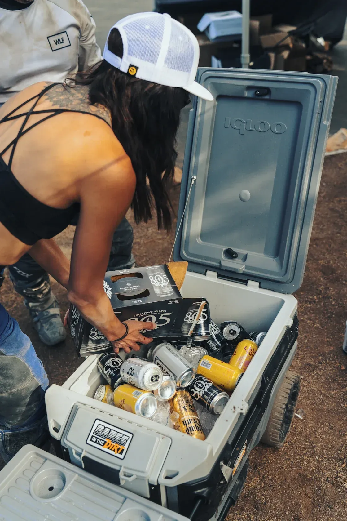 Woman reaching into a cooler filled with cans. Wearing a cap and holding a box of cans, outdoors.