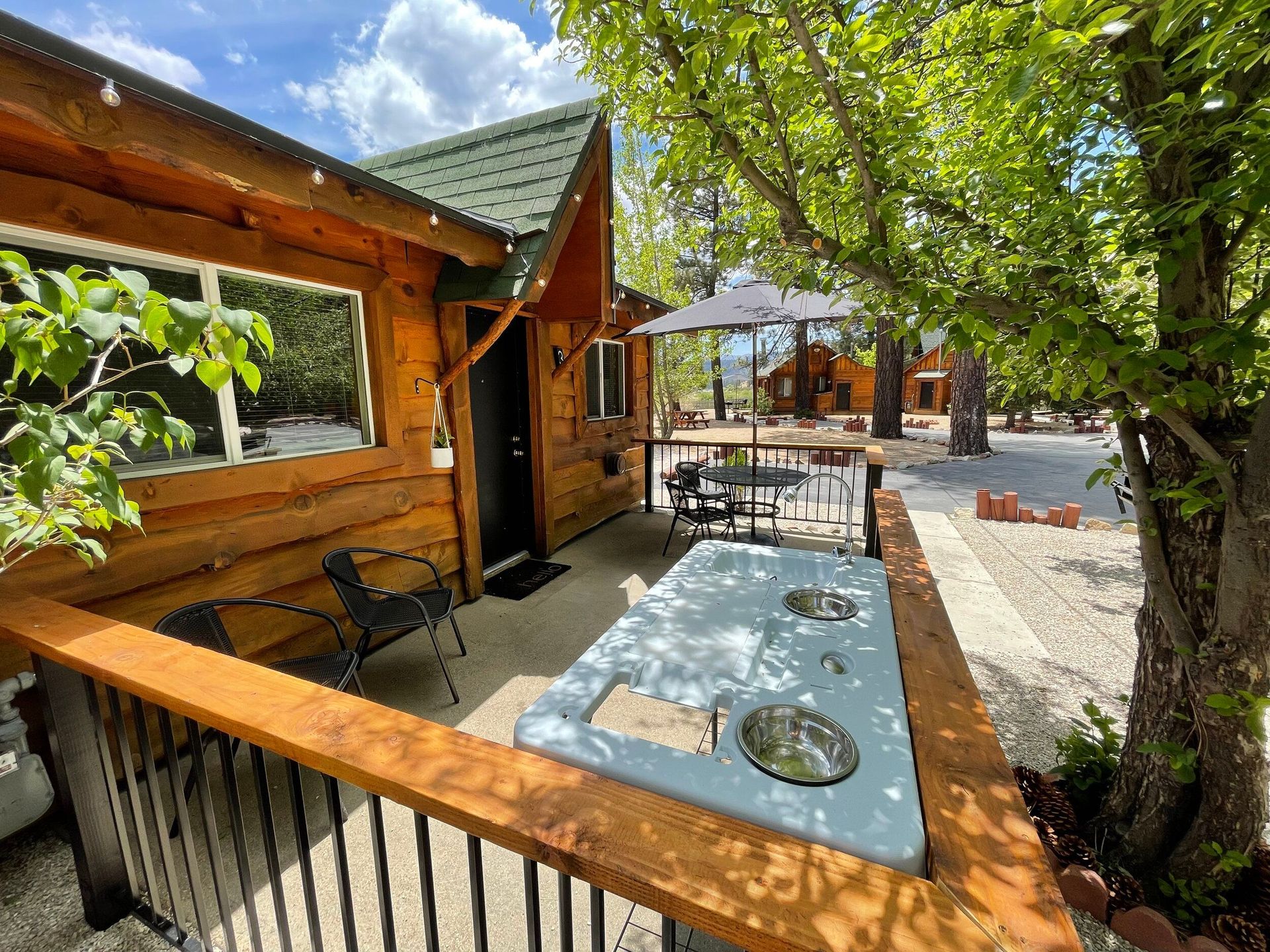 Wooden cabin exterior with porch, table, chairs, and nearby trees, sunny day.