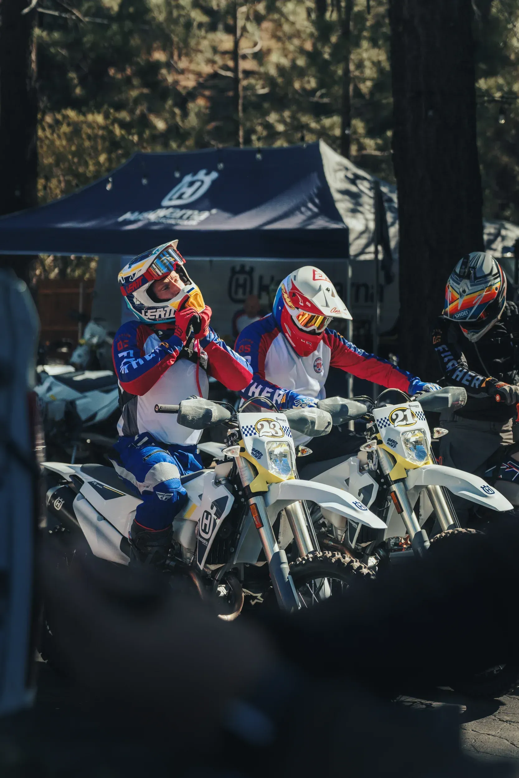 Two motocross riders on white bikes, adjusting helmets, wearing red, blue, and white gear, near Husqvarna tent.