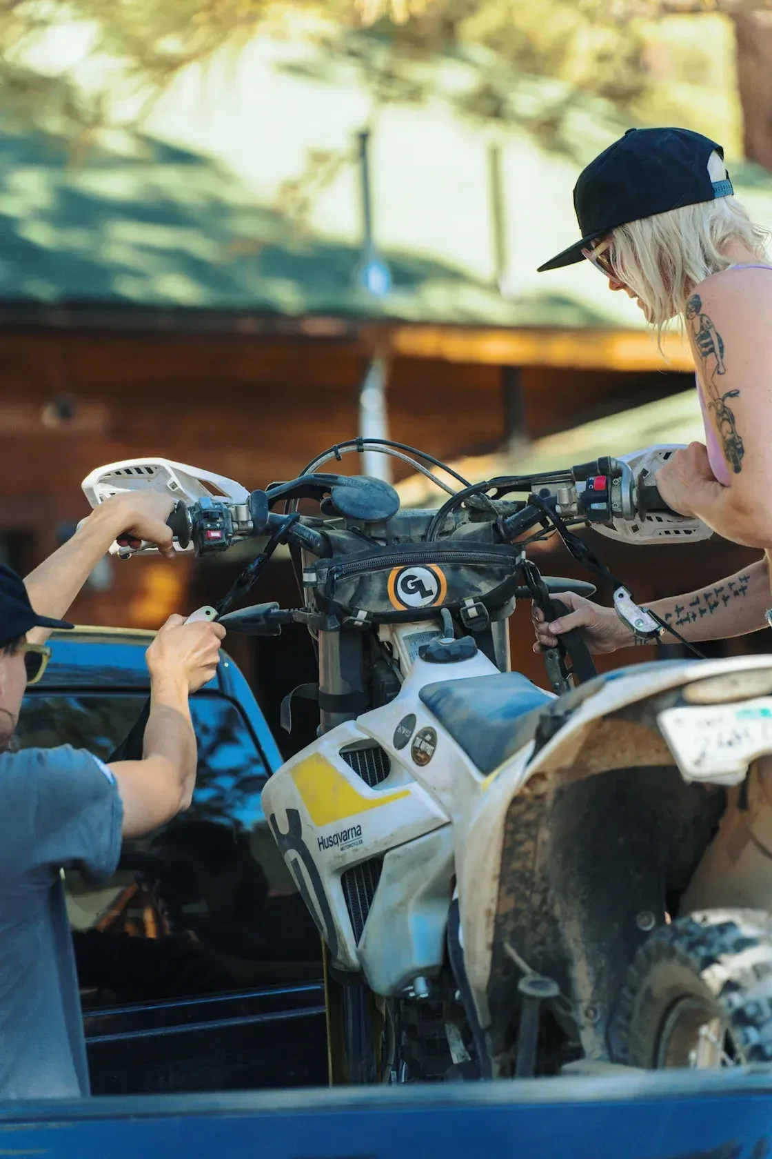 Two people loading a white motorcycle into a truck bed, in front of a wooden building.