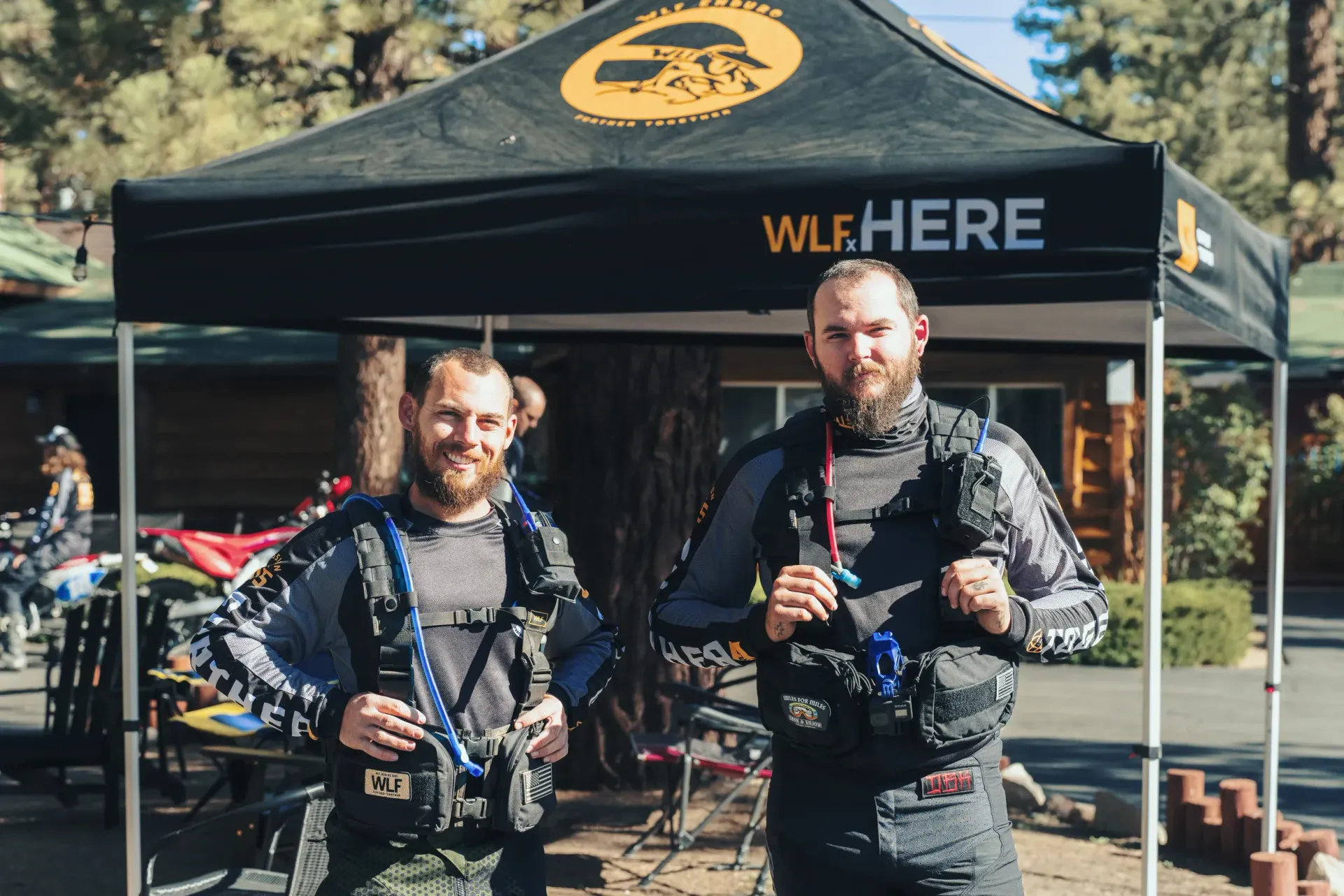 Two men with beards stand under a black tent. Both are wearing gray gear. 