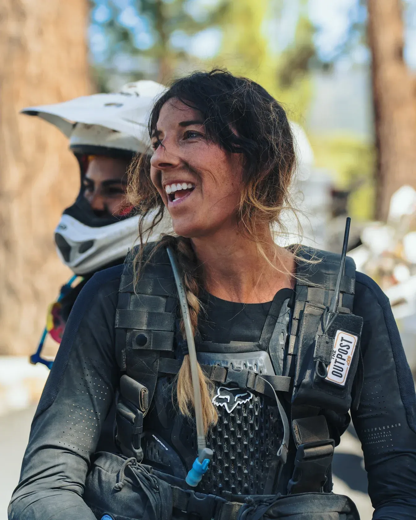 Woman in motorcycle gear smiles outdoors; another person in background.