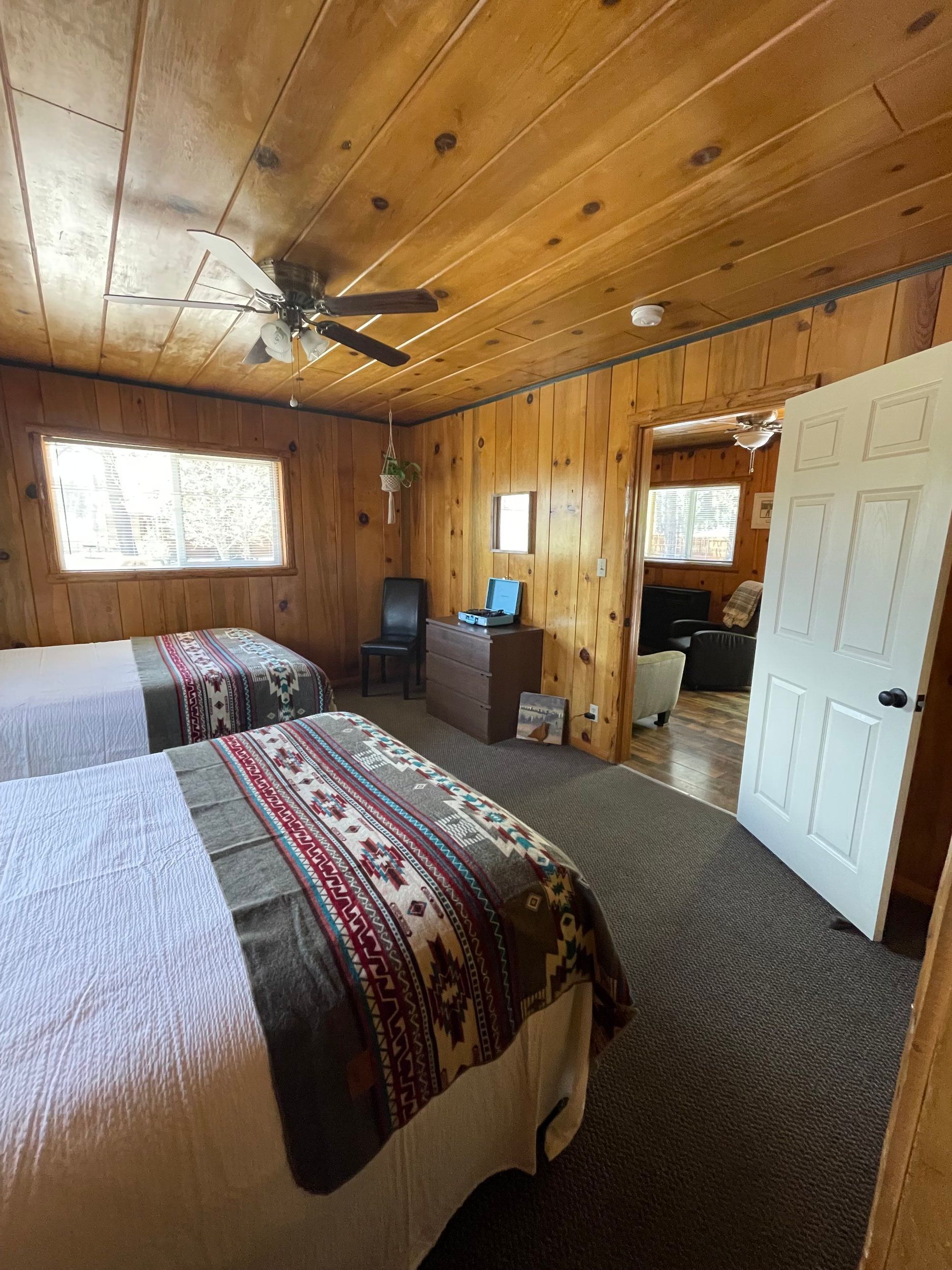A cabin bedroom with two beds, wood paneling, and a view into another room.