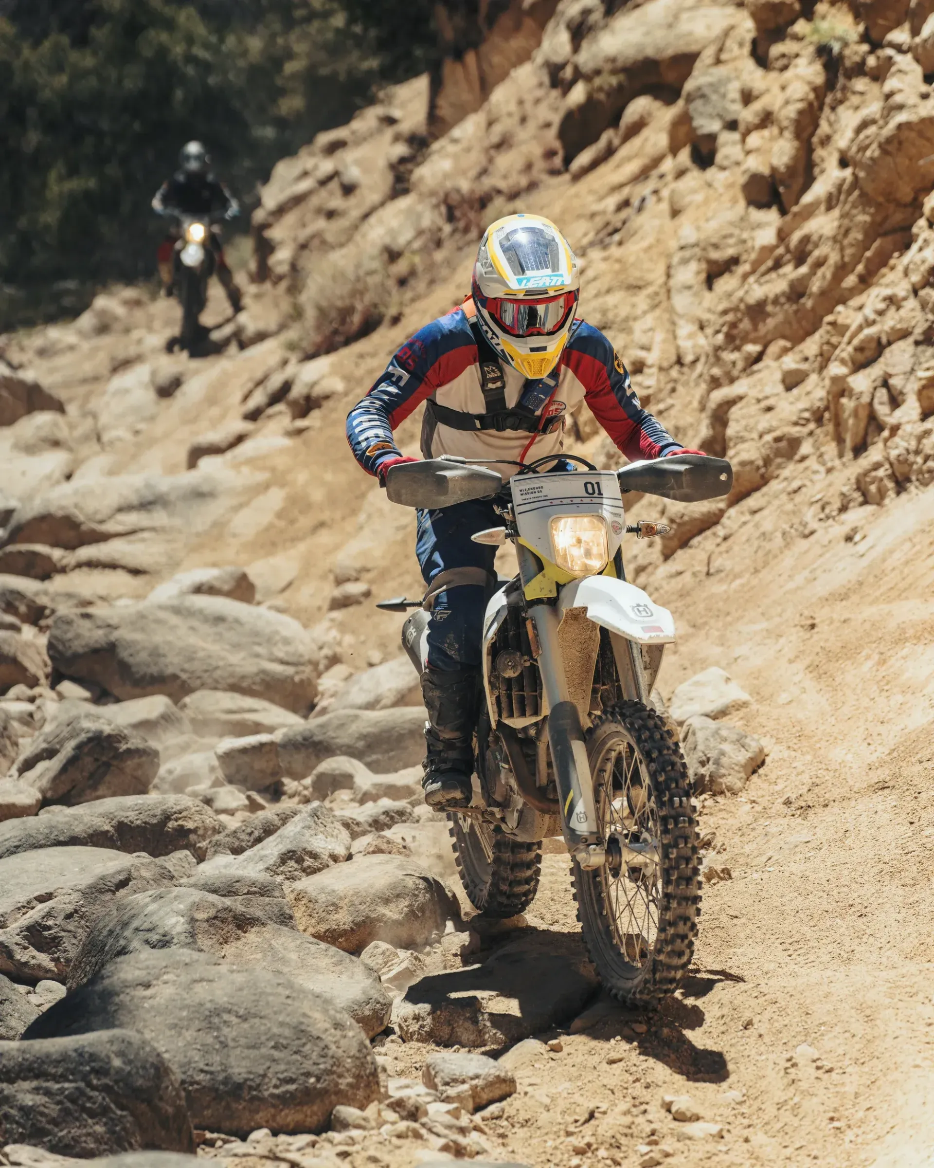 Motorcyclist riding a dirt bike up a rocky trail. Another rider follows. Sunny, daytime.