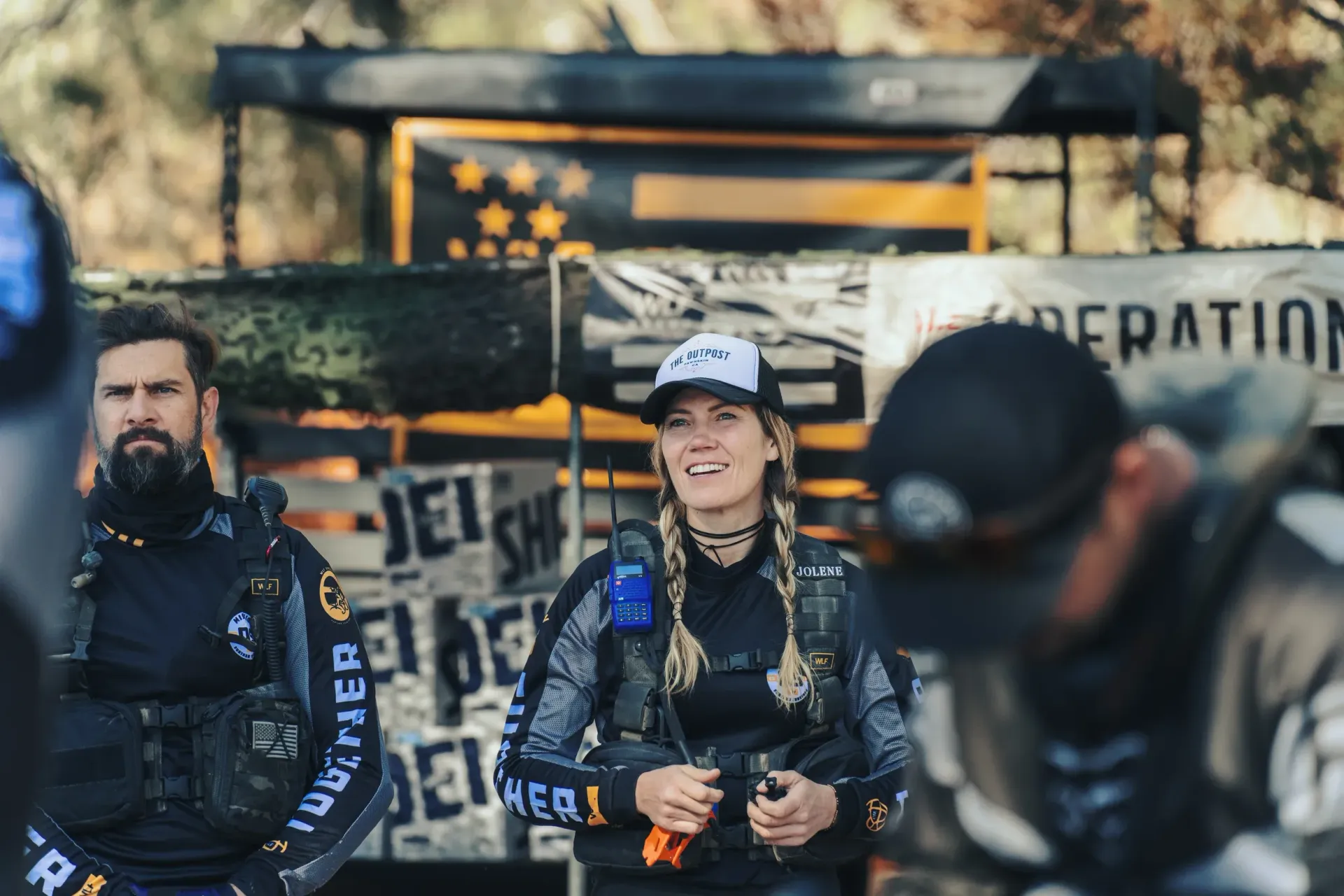 Woman smiles, flanked by people in tactical gear, outdoors; banner reads 