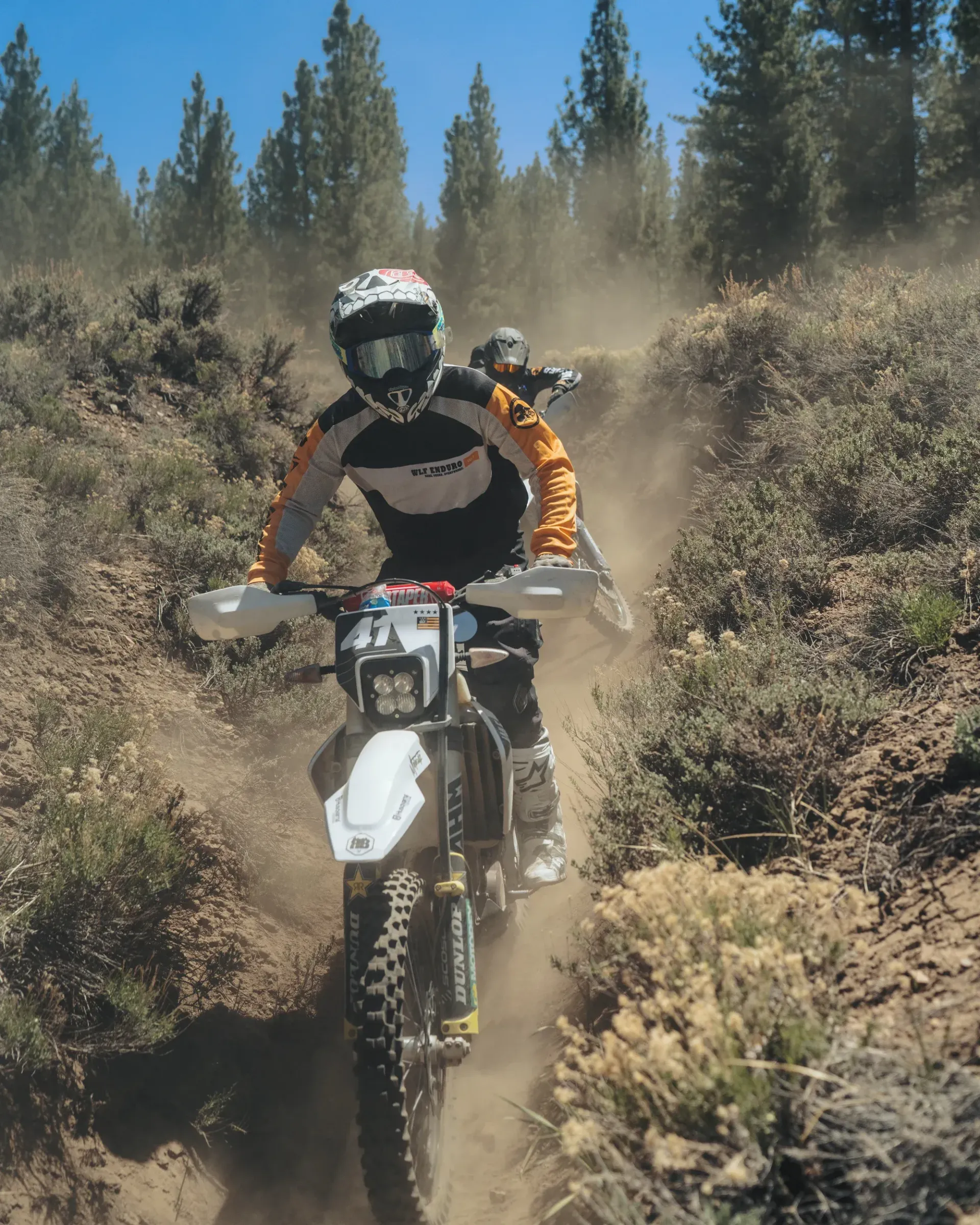 Dirt bike rider on a dusty trail, trees in the background, riding gear.