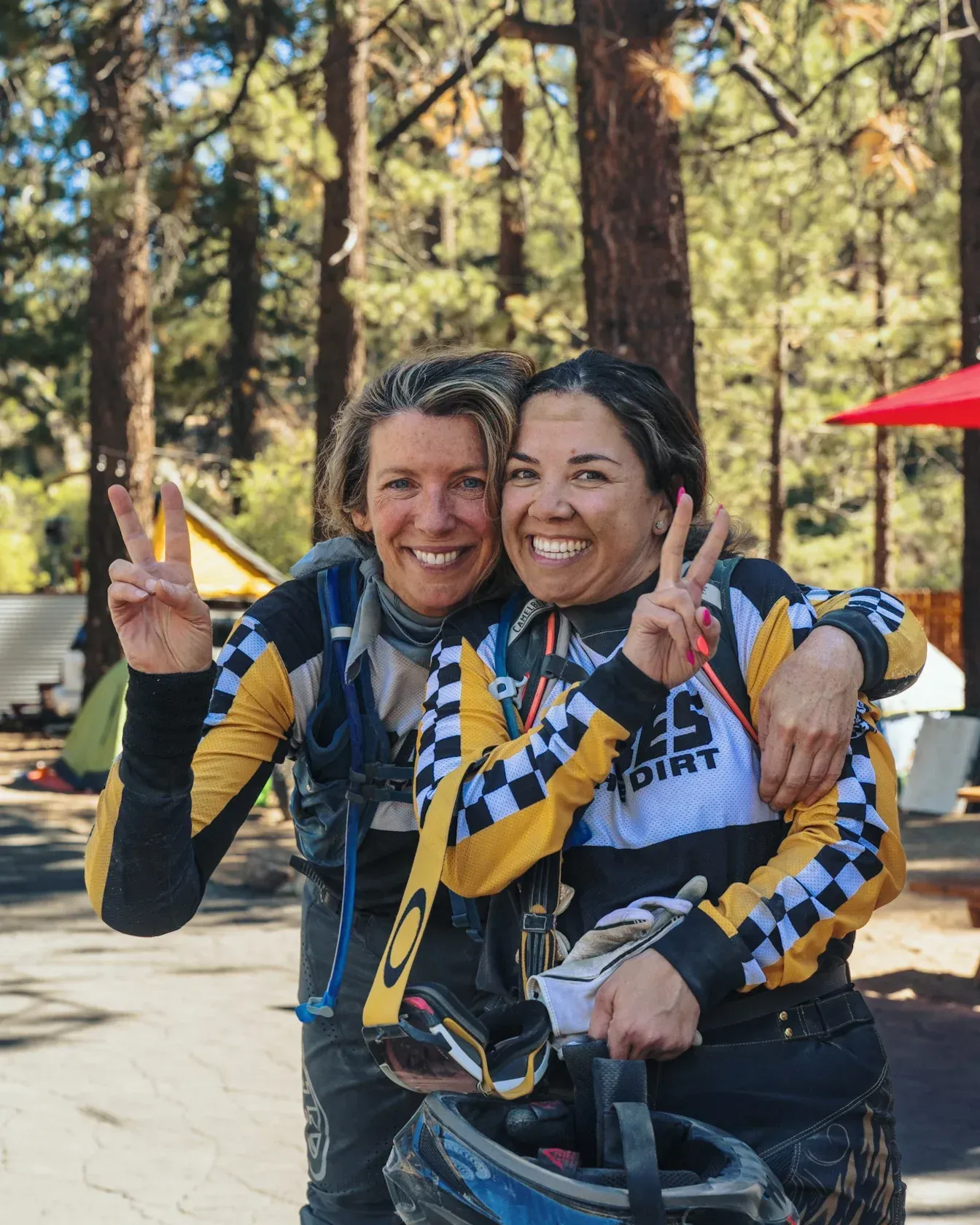 Two smiling people, wearing checkered jerseys, making peace signs. Outdoors, near trees and campsites.
