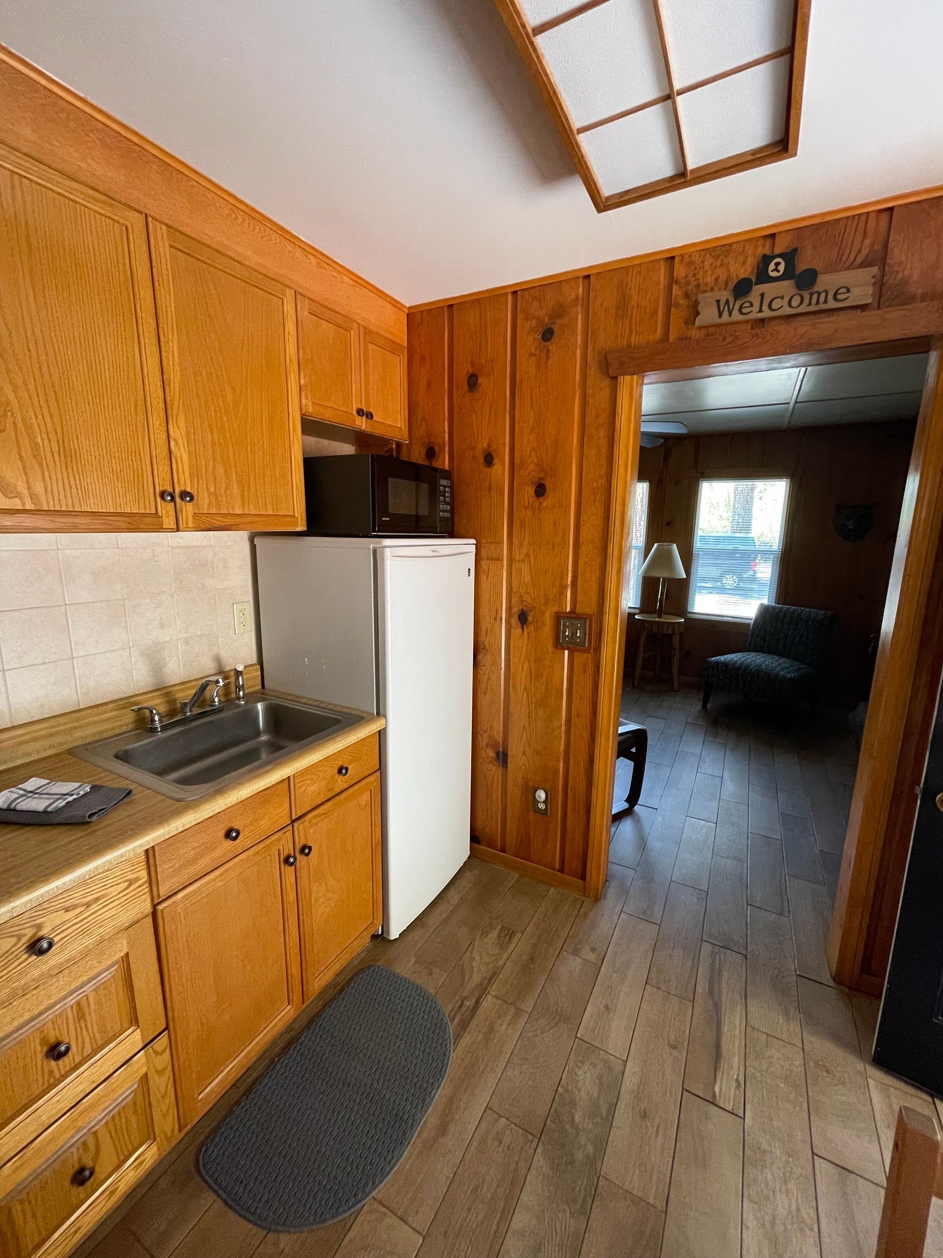 Small kitchen with wood cabinets, sink, mini-fridge, and view into living area.