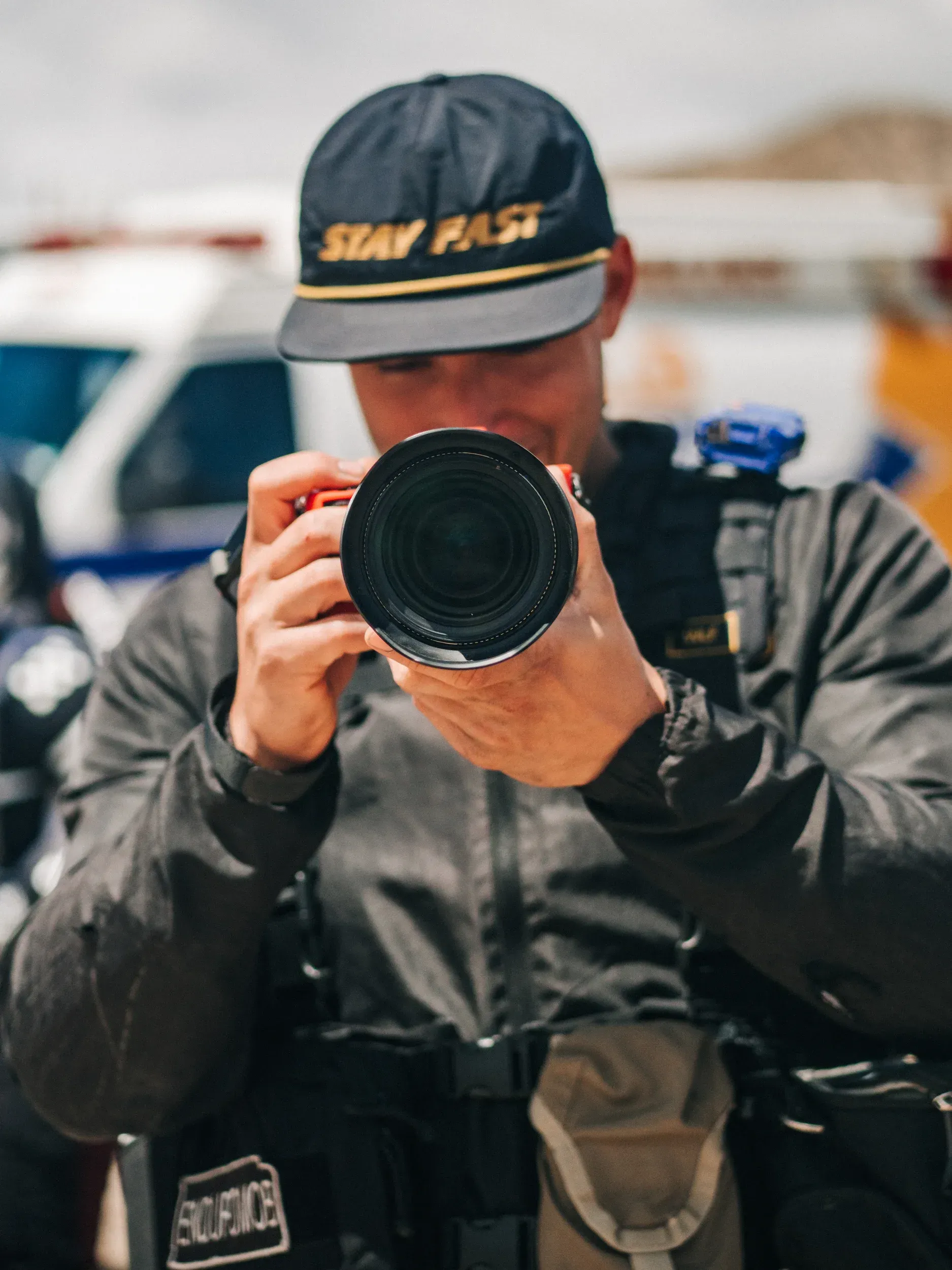 Person wearing a hat and black vest, holding a red camera and looking through the lens.