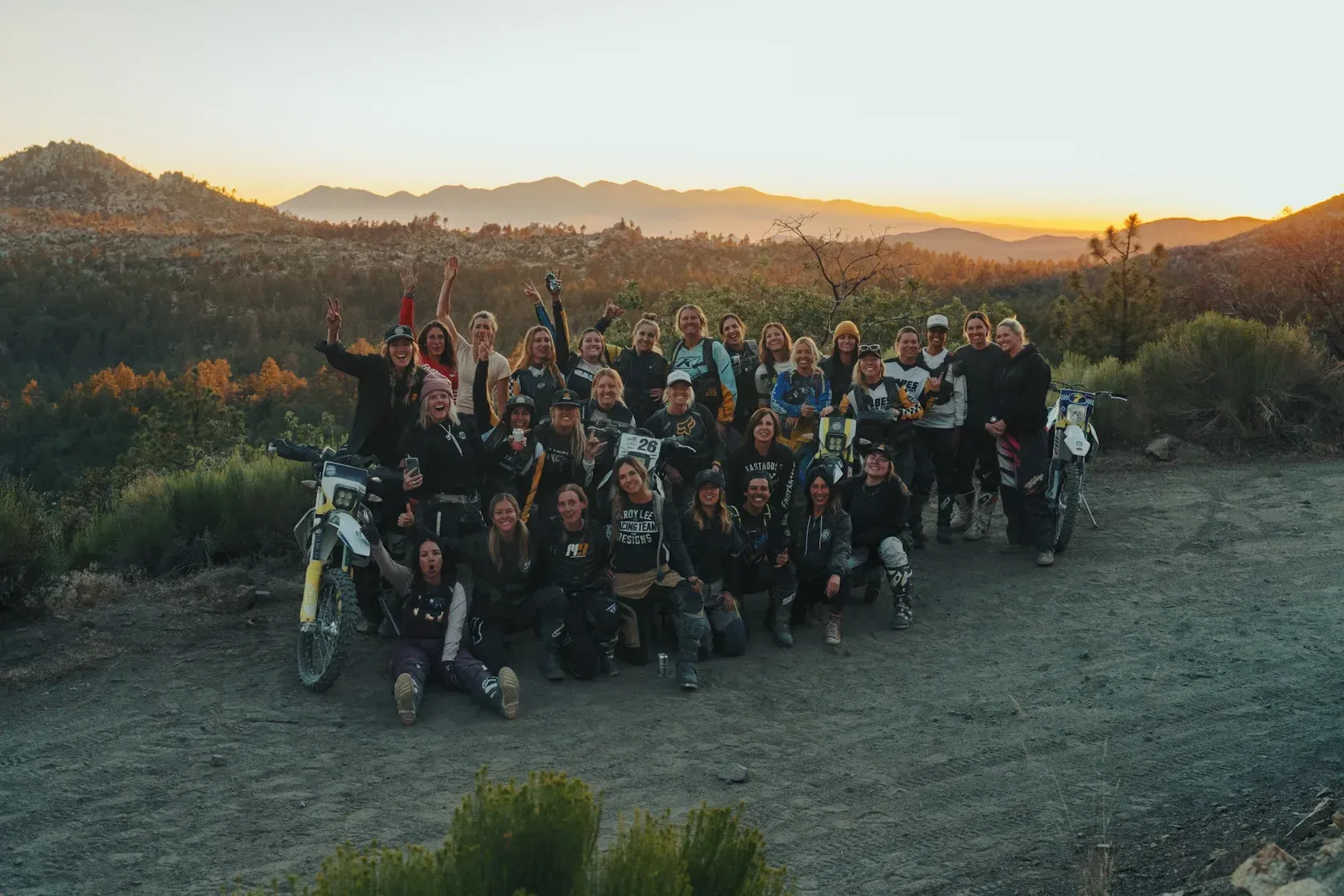 Group of people on dirt bikes posing at sunset in a mountainous area. Hands raised, smiling.
