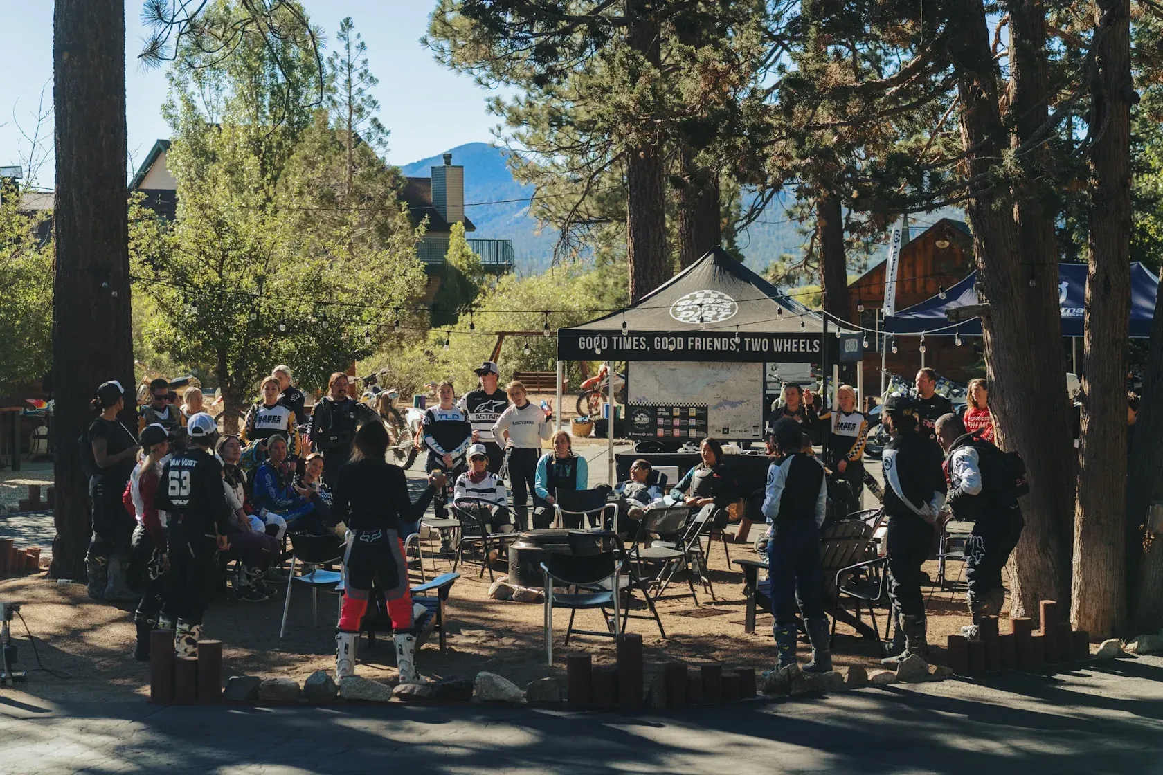 Group of people gathered outdoors, possibly at an event, near a sign; trees and mountains visible.