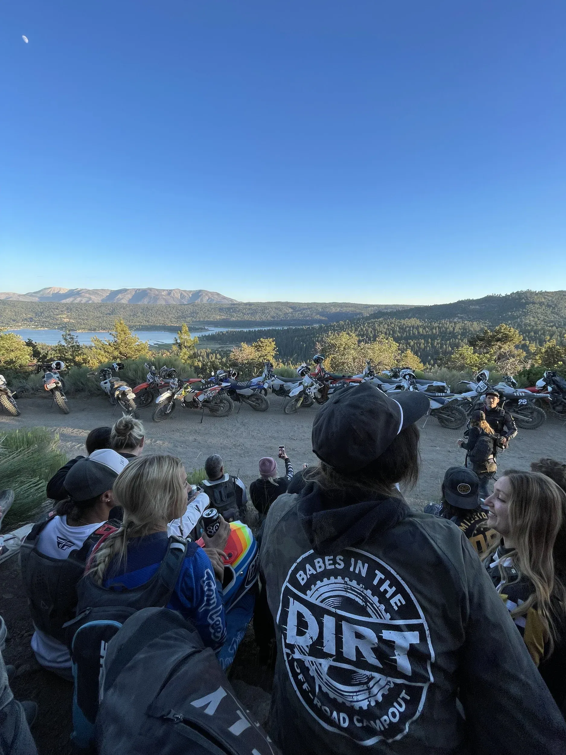 People watch motorcycles on a hillside overlooking a lake under a blue sky; one person wears a 