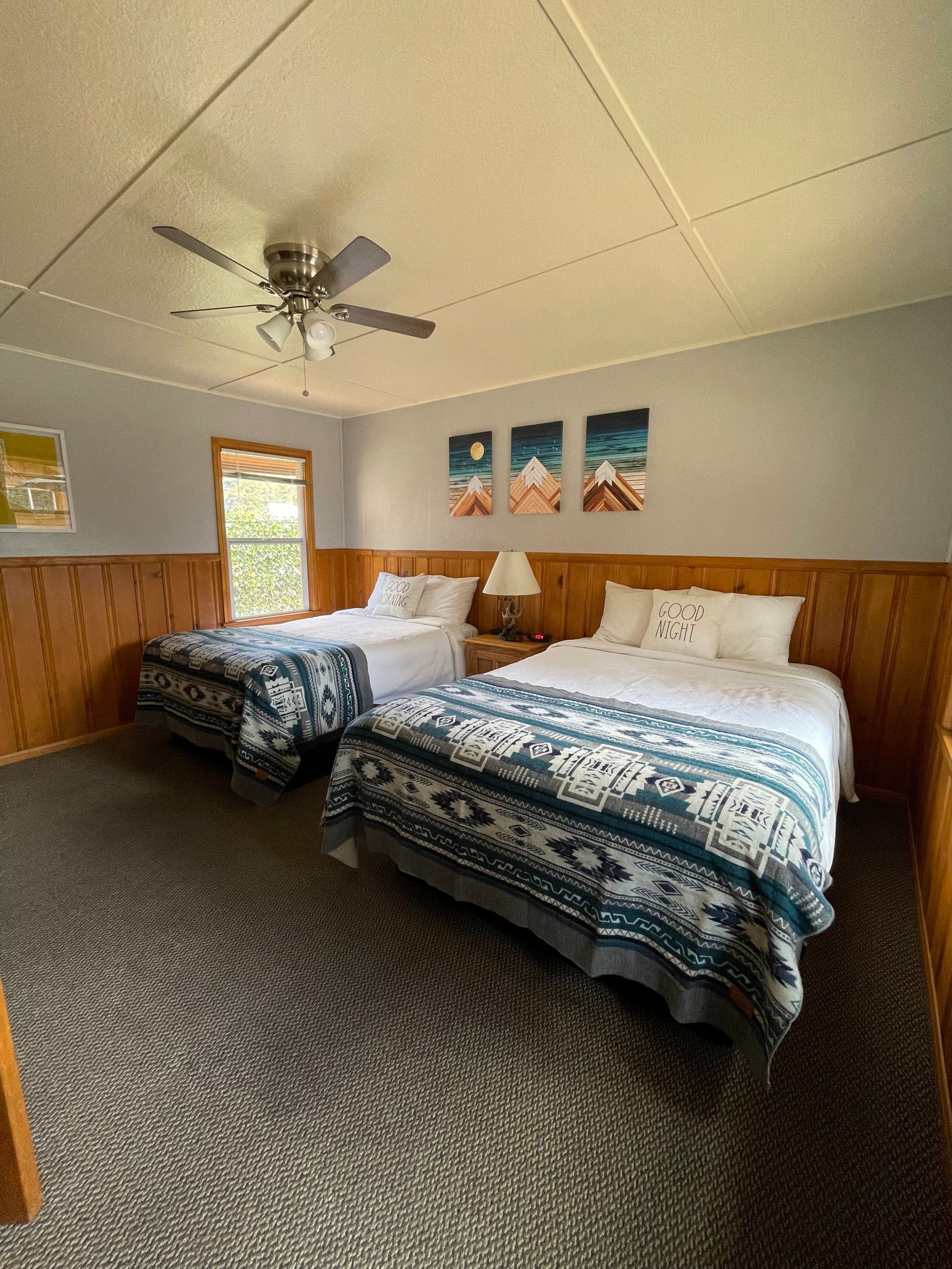 Bedroom with two beds, gray carpet, wood-paneled walls, ceiling fan, and mountain art.