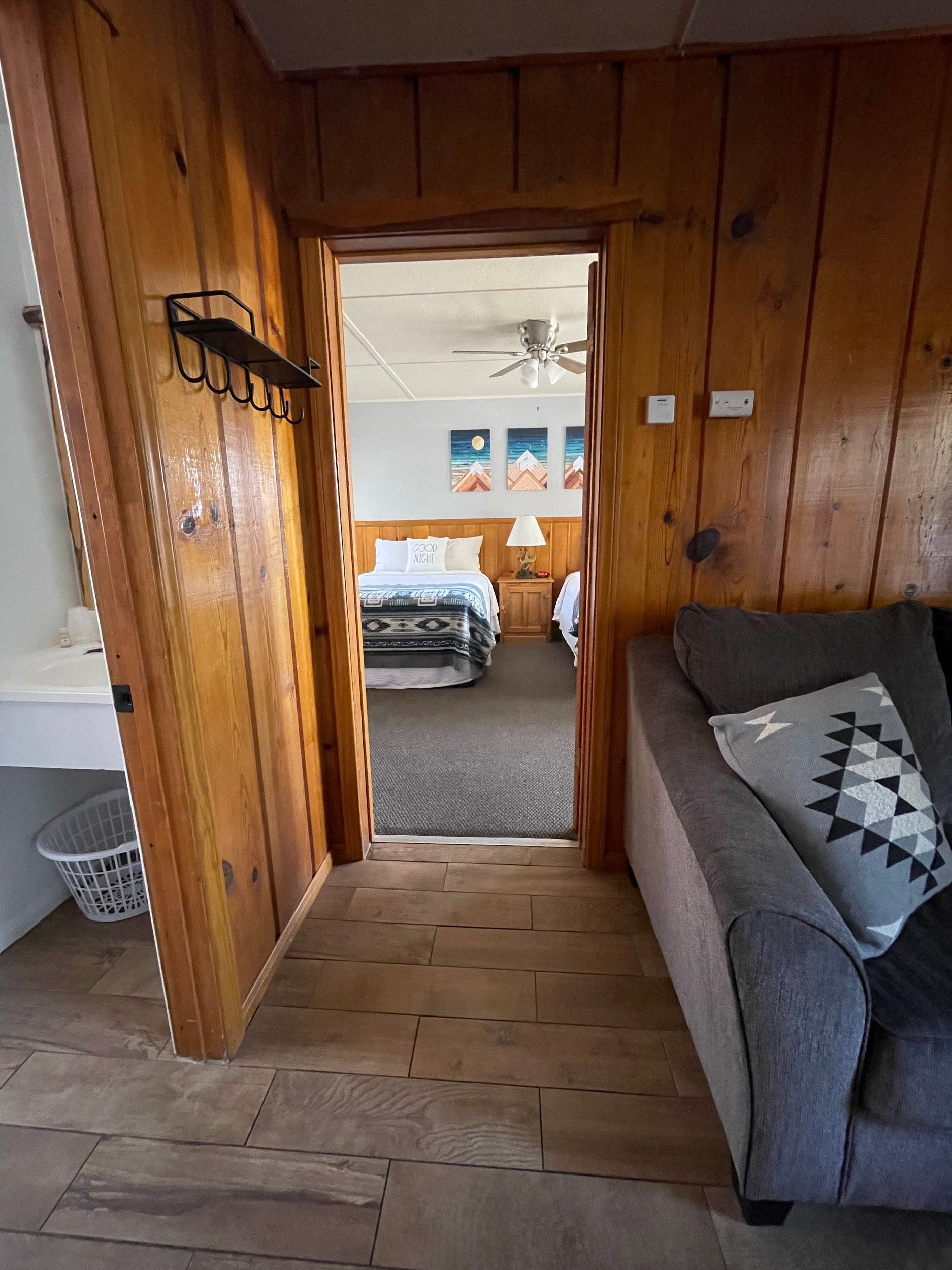 View from doorway into a bedroom. Wooden paneling surrounds doorway, leading to a bed. Gray sofa to the right.