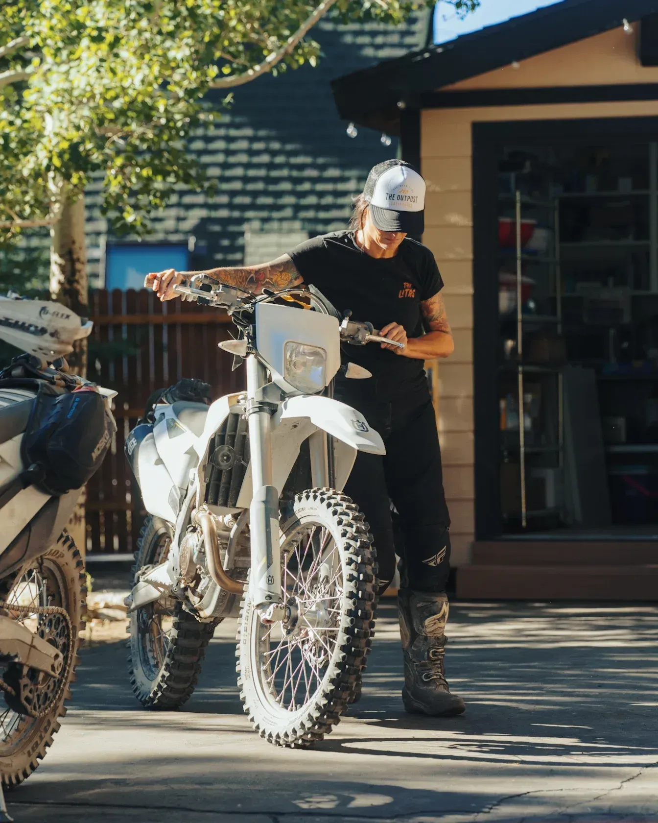 Person adjusting controls on a white dirt bike outdoors. Wearing a black outfit, boots, and a hat.