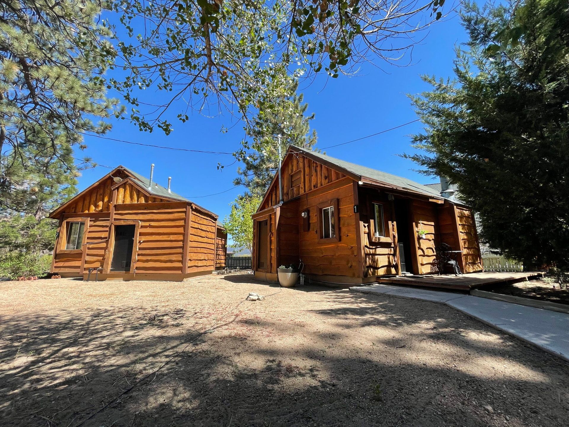 Two rustic wooden cabins under a blue sky, surrounded by trees and a gravel yard.