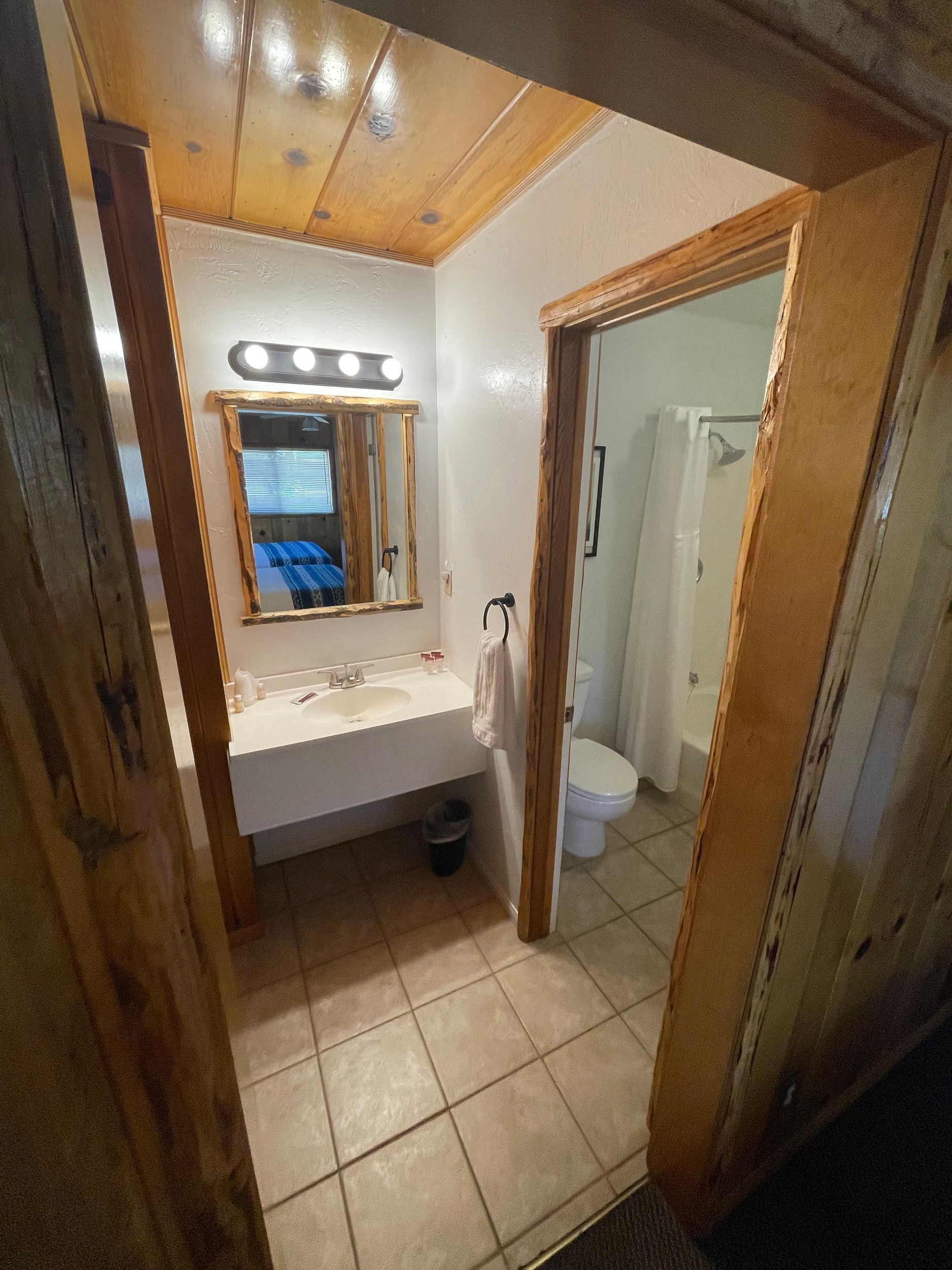 Bathroom interior with a vanity, mirror, toilet, and shower. Tan tile floor, light walls, and wooden trim.