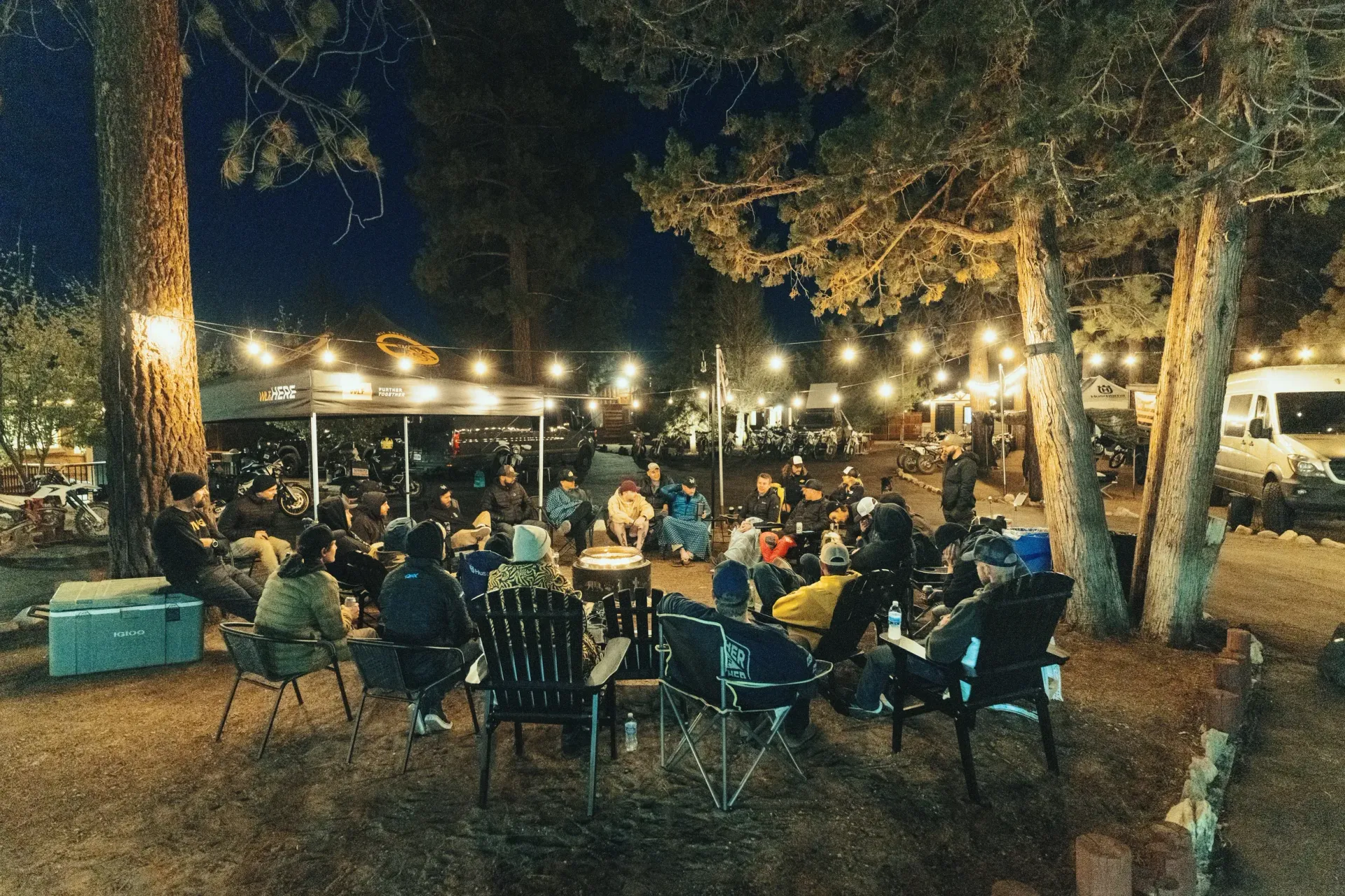 People gathered around a fire pit at night, strung with lights under trees.