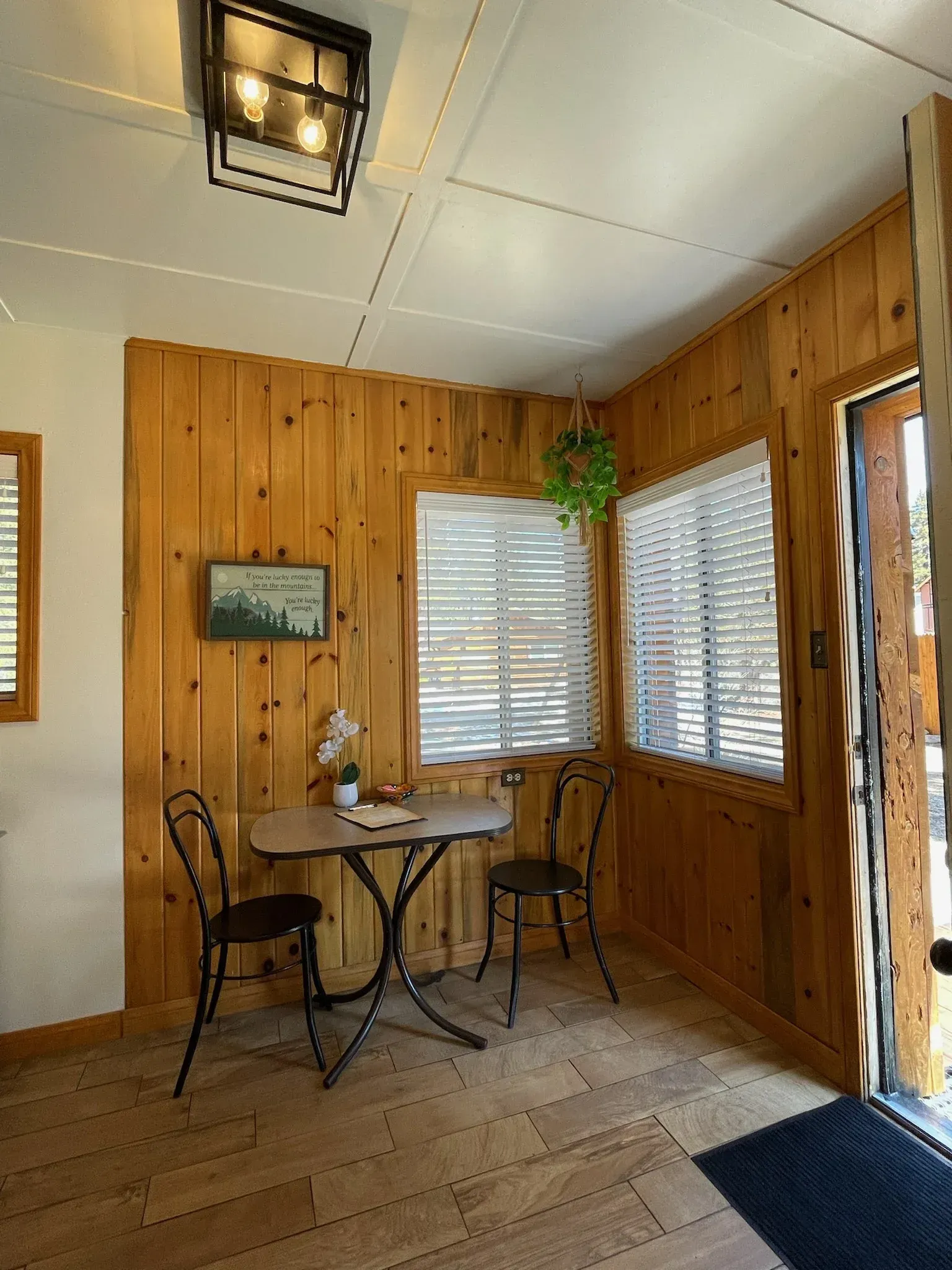 Cozy nook with wood-paneled walls, table, two chairs, and two windows with blinds. Decorative hanging plant and a black ceiling light.