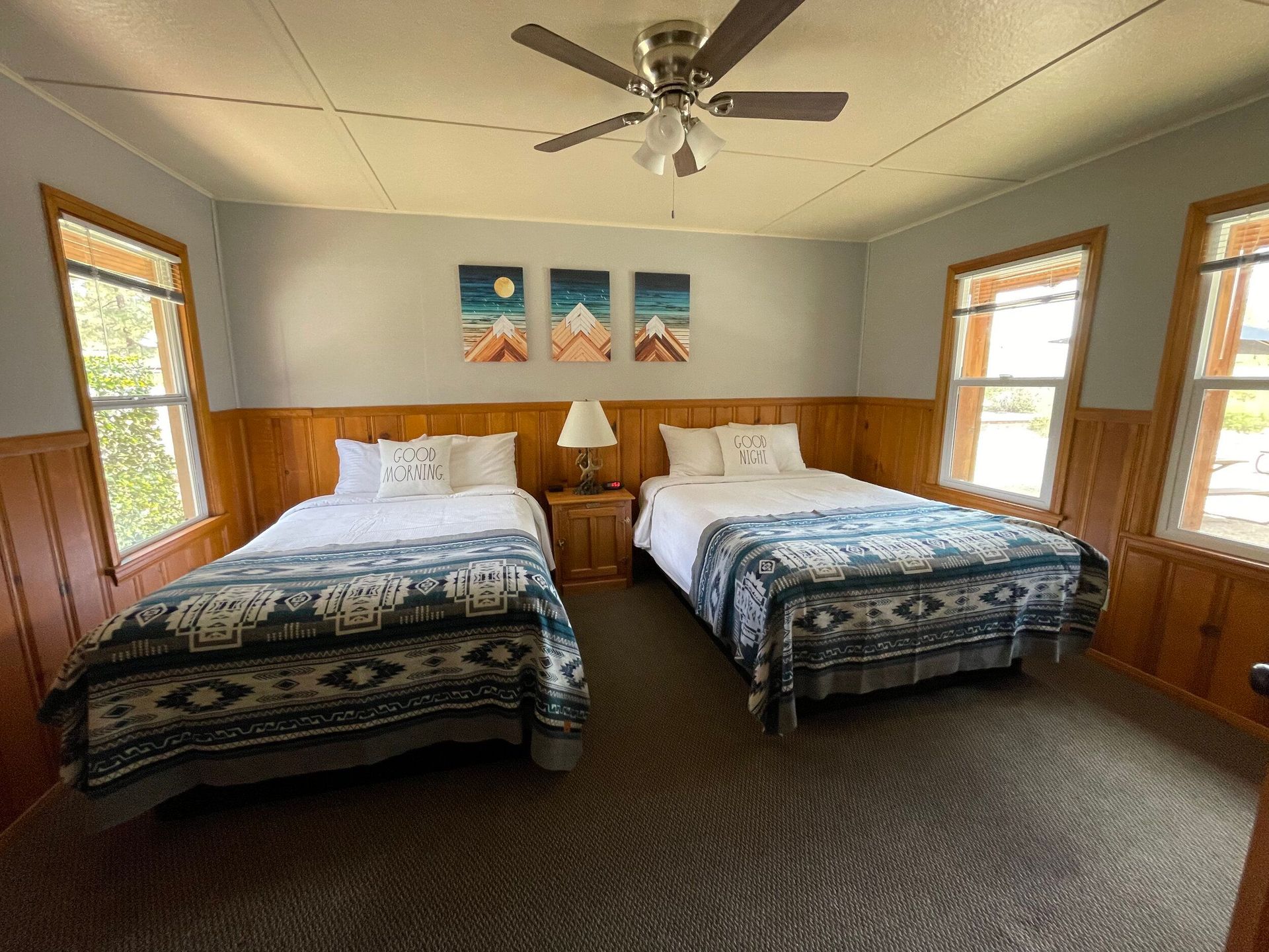 Bedroom with two beds, wooden accents, windows, artwork, and a ceiling fan.