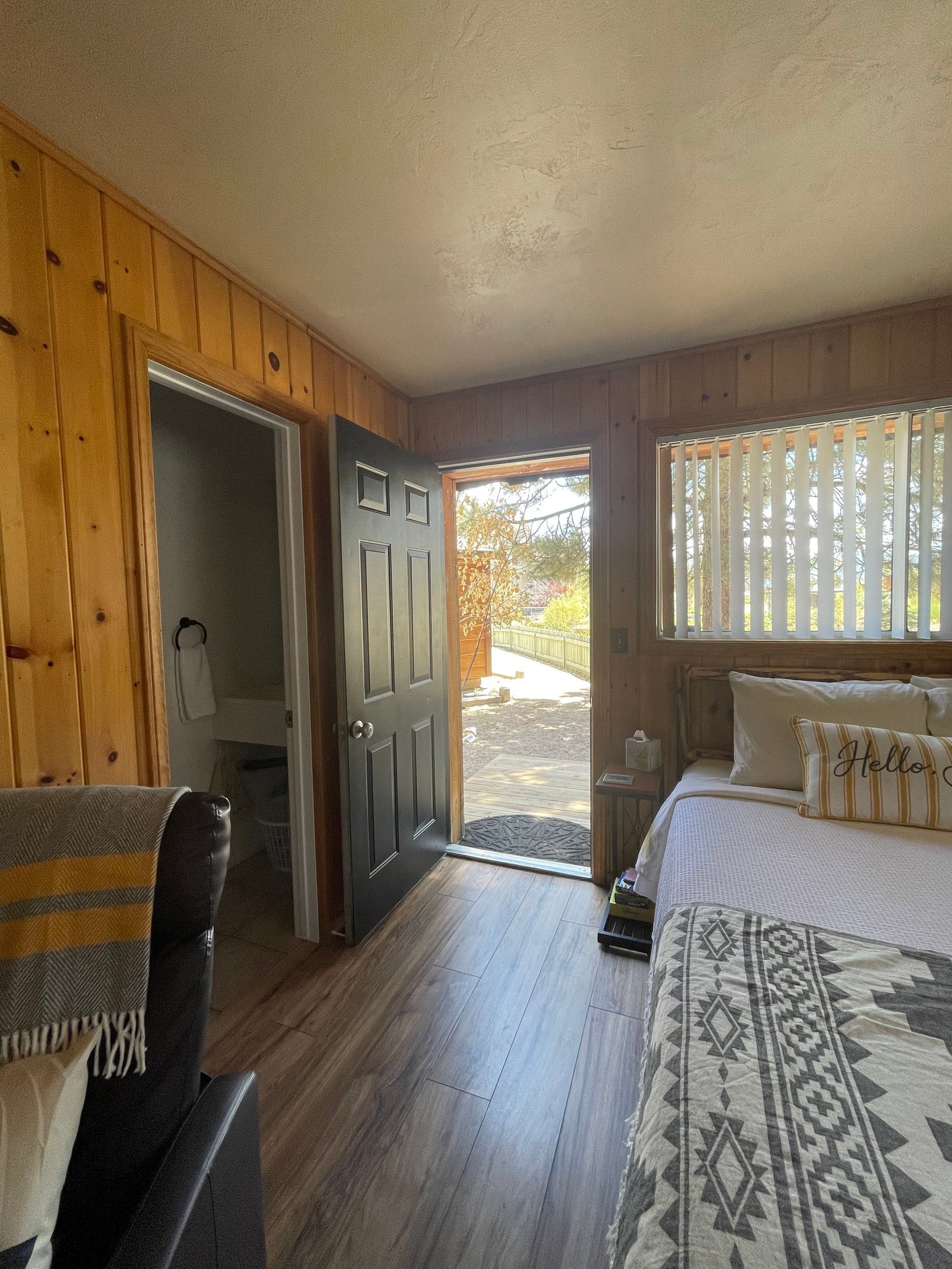 Bedroom with wood paneling, open black door, bed with patterned cover, bathroom door on the left, and a window with blinds on the right.
