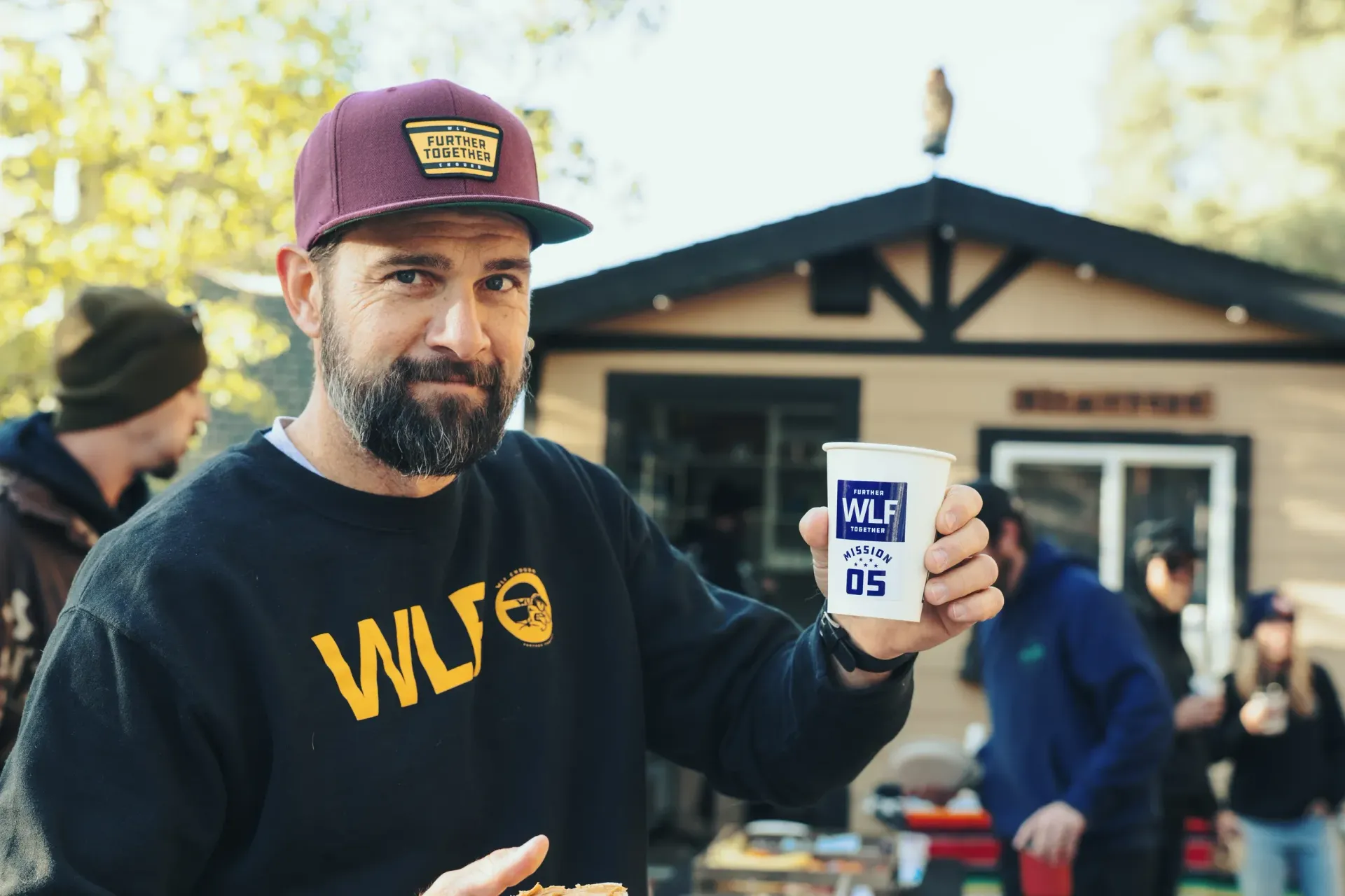 Man holding a cup, smiling, wearing a baseball cap and sweater, standing outside a small building.