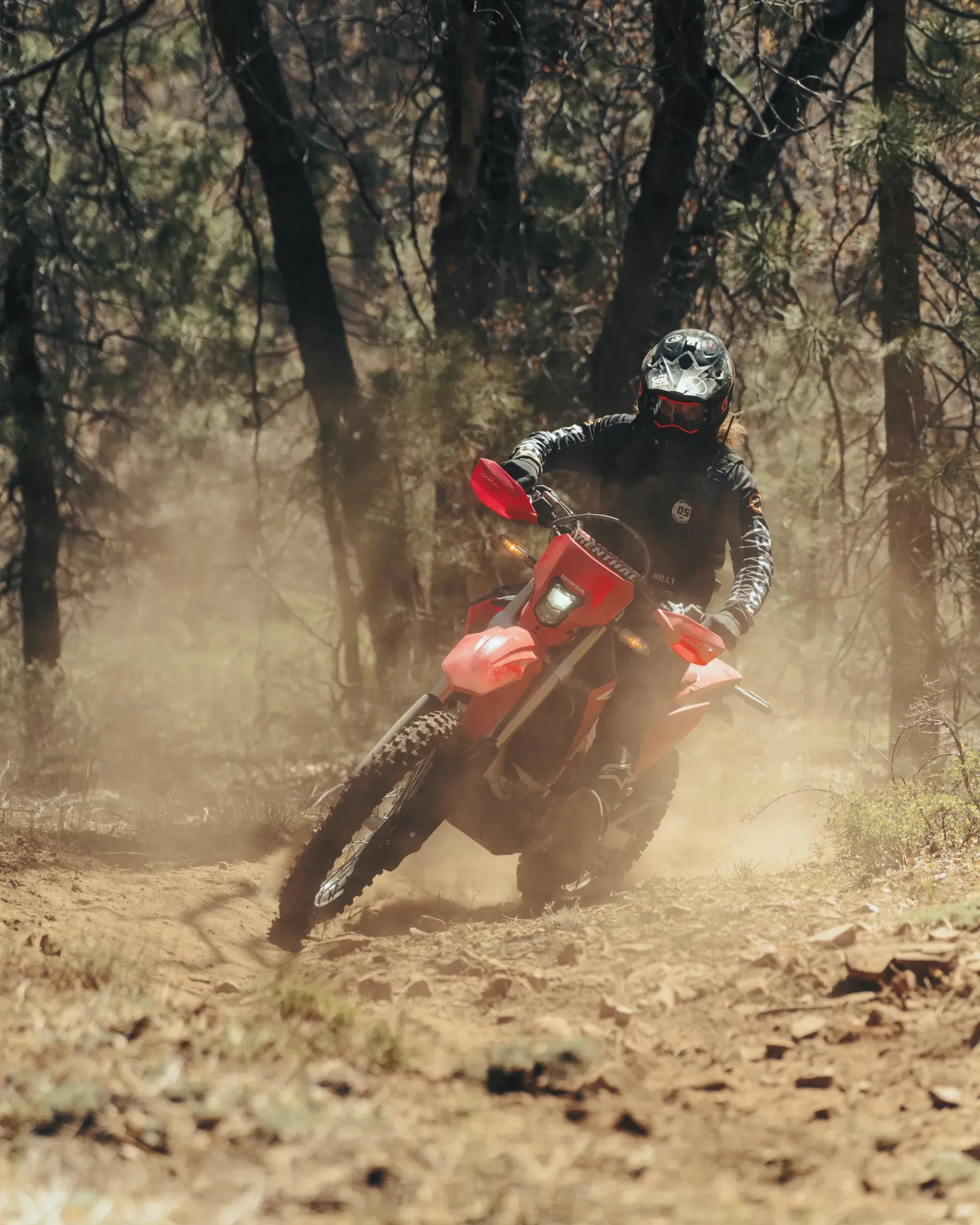 Dirt bike rider leans into a turn on a dirt trail, kicking up dust in a forest.