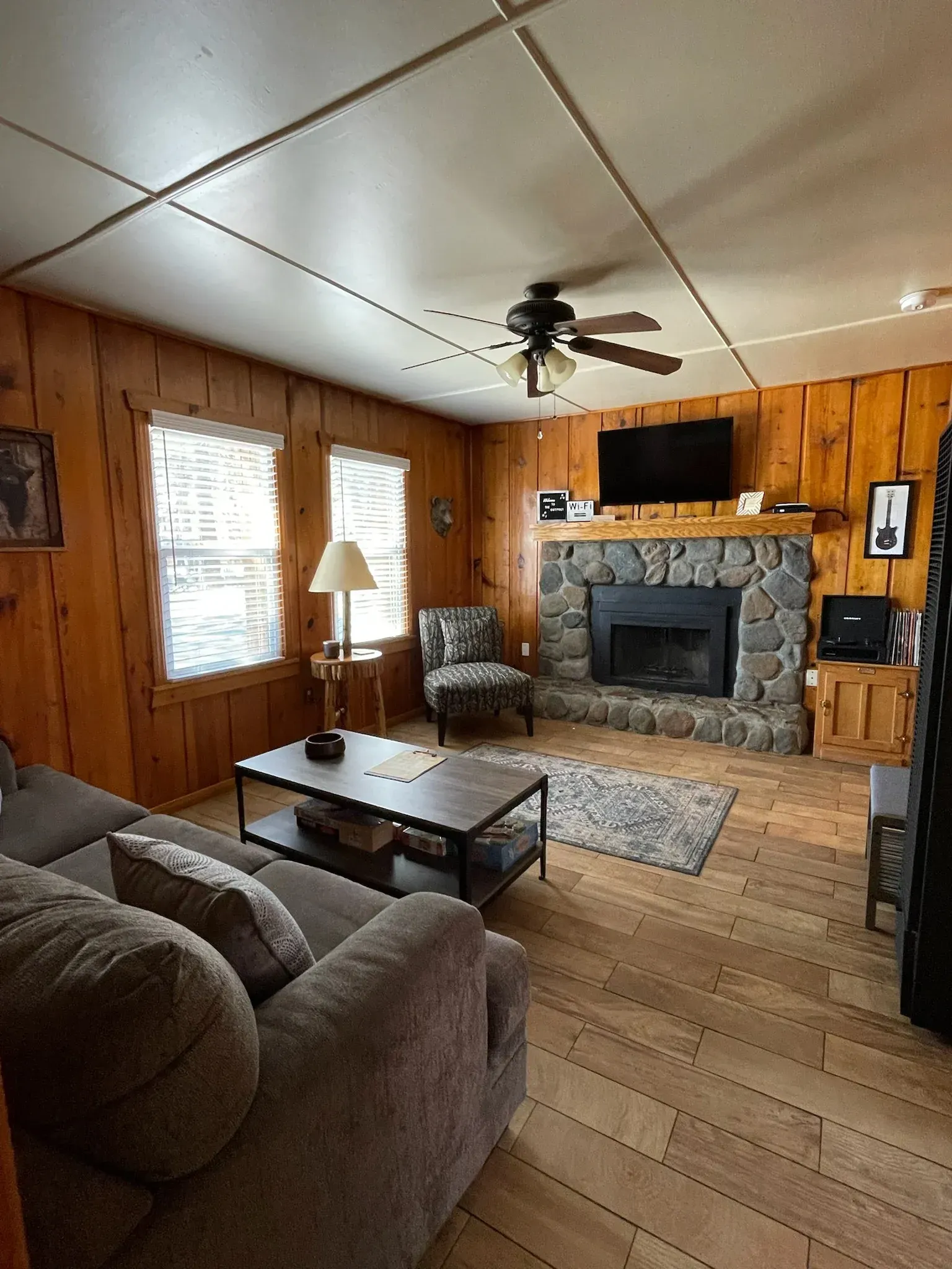 Cozy living room with stone fireplace, wood paneling, gray couch, and TV.