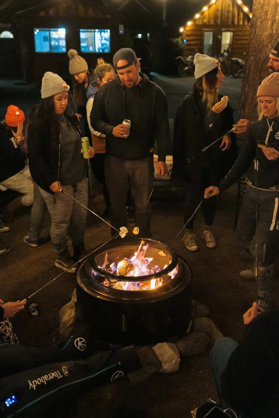 People roasting marshmallows over a fire pit outdoors at night. Several wear hats.