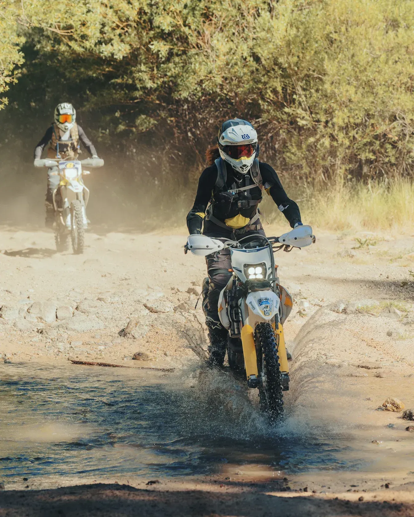 Two dirt bikes riding through water on a dirt road, kicking up water and dust.