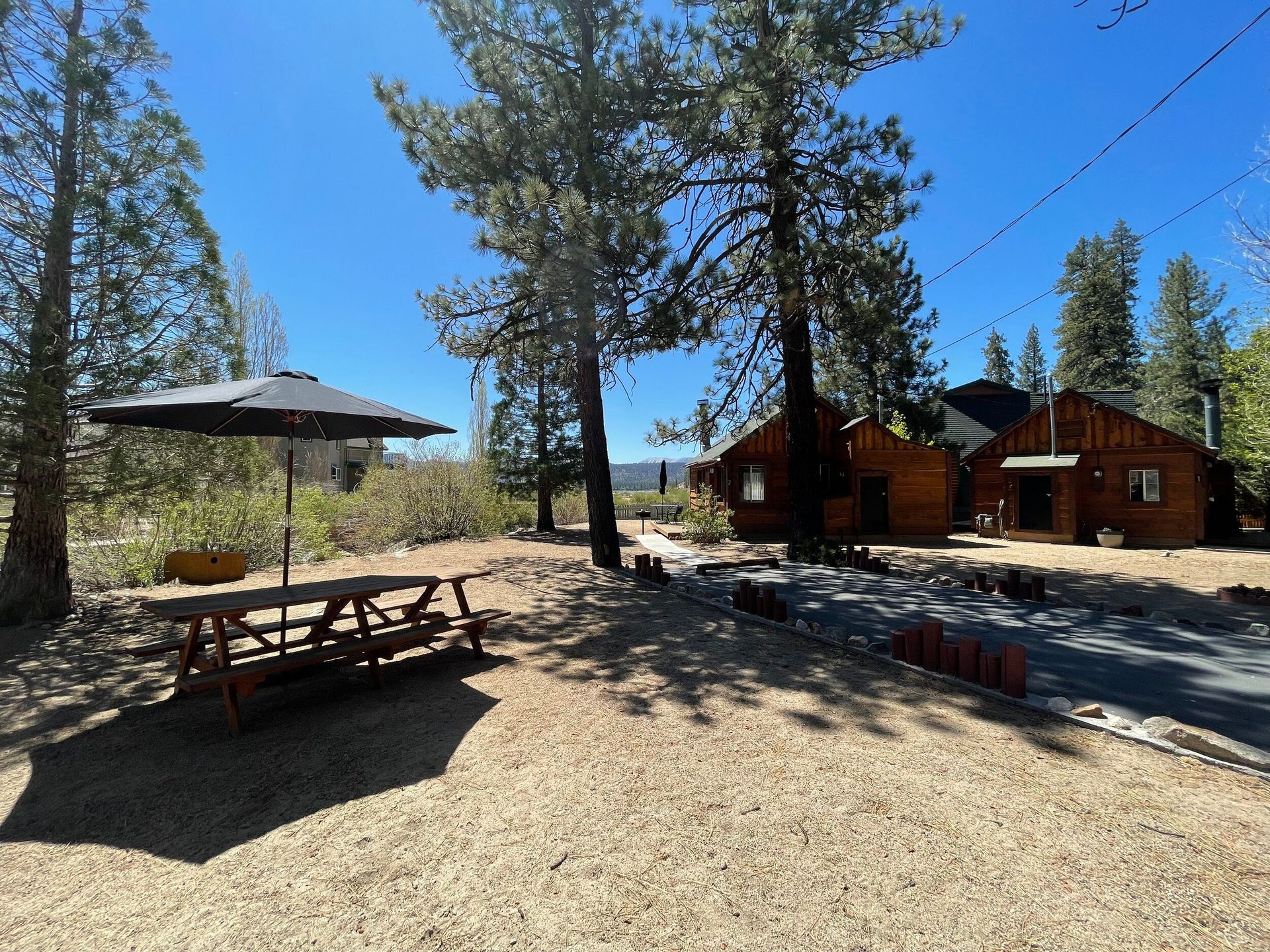 Wooden cabin and picnic table under an umbrella on a gravel lot, trees in background under a blue sky.