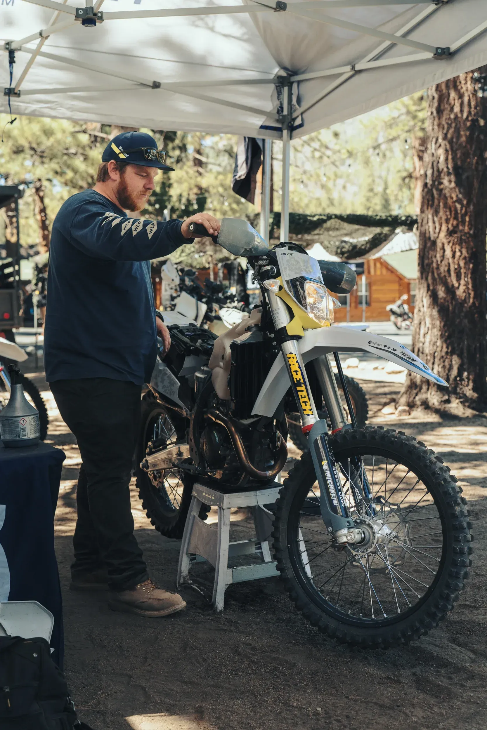 Man working on a motorcycle under a canopy. Bike is gray, white, and yellow. Man wears a blue shirt and hat. Outdoors.
