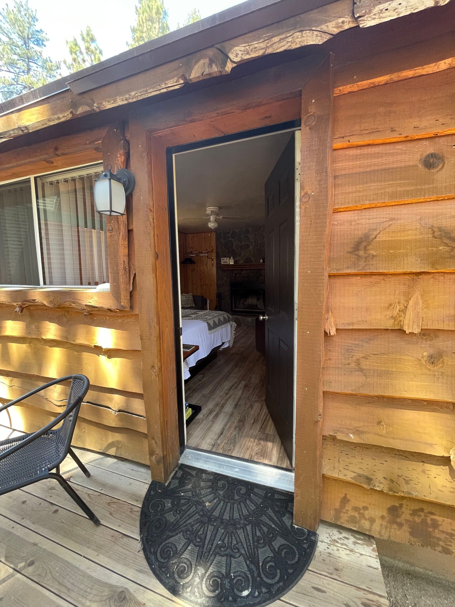 Doorway of a wooden cabin, open to the interior with bed and fireplace. Exterior has a chair and doormat.