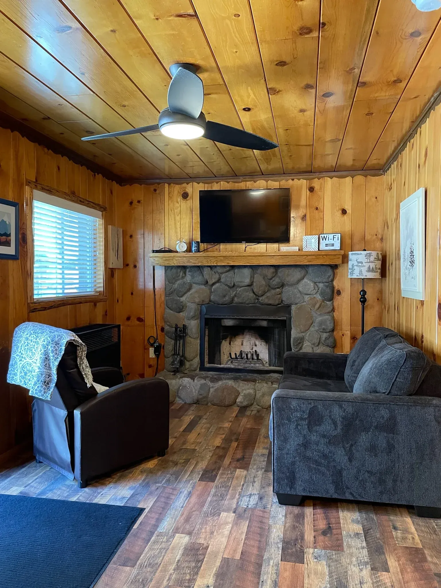Cozy cabin living room with wooden walls, fireplace, sofa, recliner, TV, and ceiling fan.